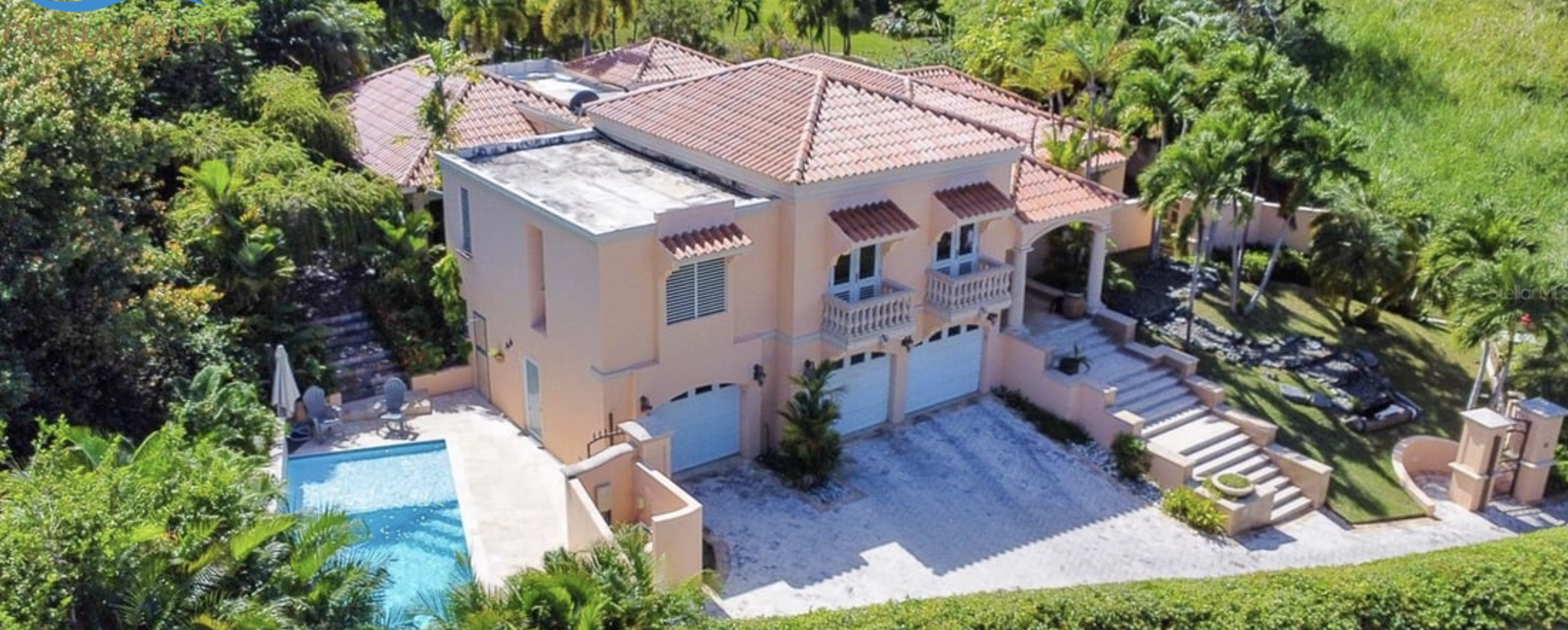 Aerial view of a two-story house with a red-tiled roof, pink exterior walls, small balconies, surrounded by lush greenery, trees, and a backyard with a swimming pool and patio.