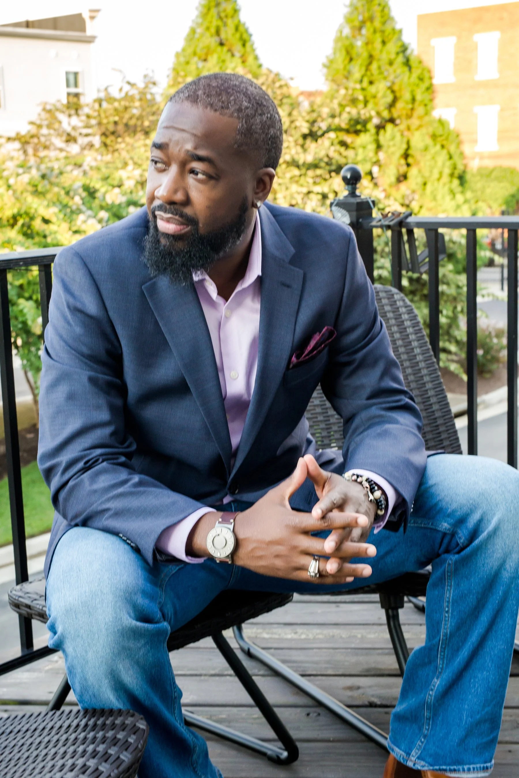 A man with a beard wearing a navy blazer, pink shirt, and jeans sitting on a balcony with greenery and buildings in the background.