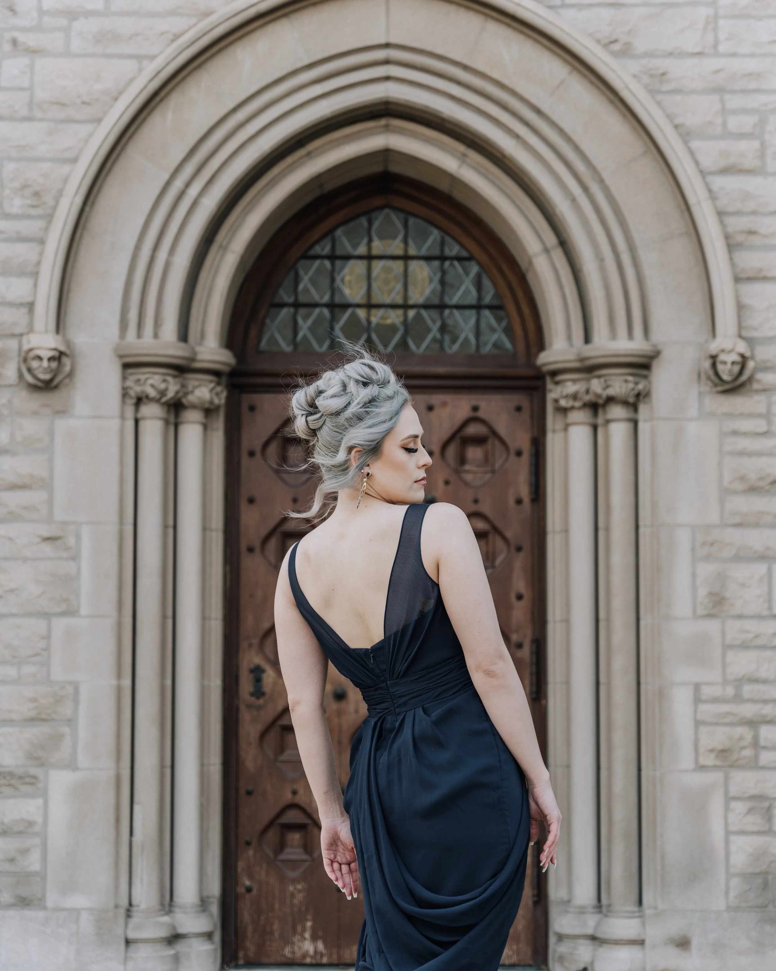 Side profile of a woman with silver hair styled in an elegant updo, wearing earrings and a dark, sleeveless dress, standing in front of a historic stone building with an arched wooden door and Gothic architectural details.