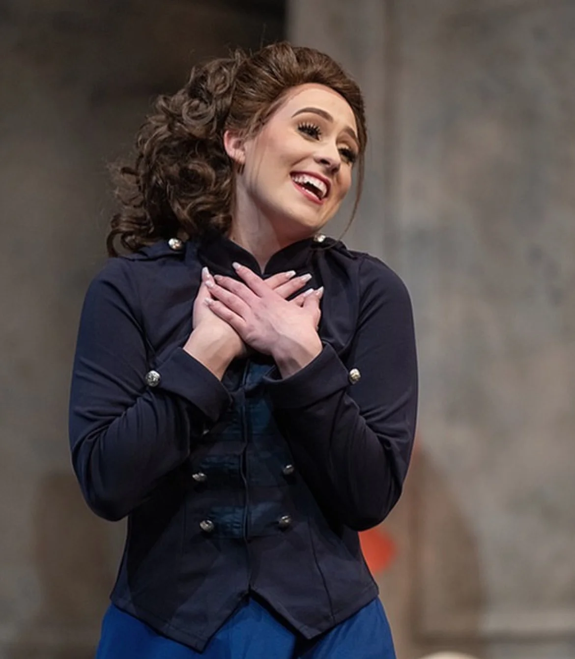 A woman with curly brown hair smiling and holding her hands on her chest, wearing a dark navy coat with pearl buttons.