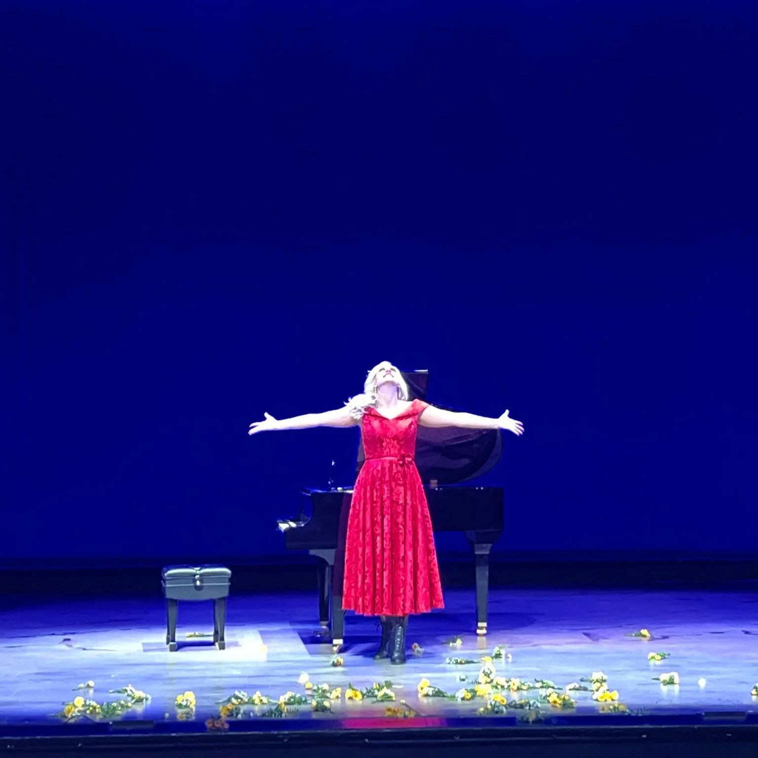 A woman in a red dress with long, blonde hair stands on stage with her arms outstretched, facing upward, in front of a blue background. There is a black grand piano behind her, and yellow flowers are scattered on the stage floor.