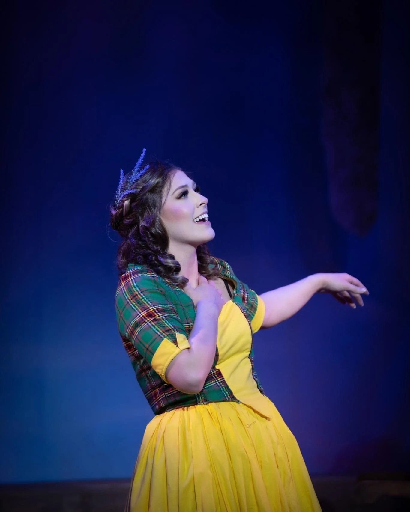 A woman on stage wearing a plaid top with yellow sleeves and a yellow skirt, smiling and gesturing, with a dark background and a lavender crown on her head.
