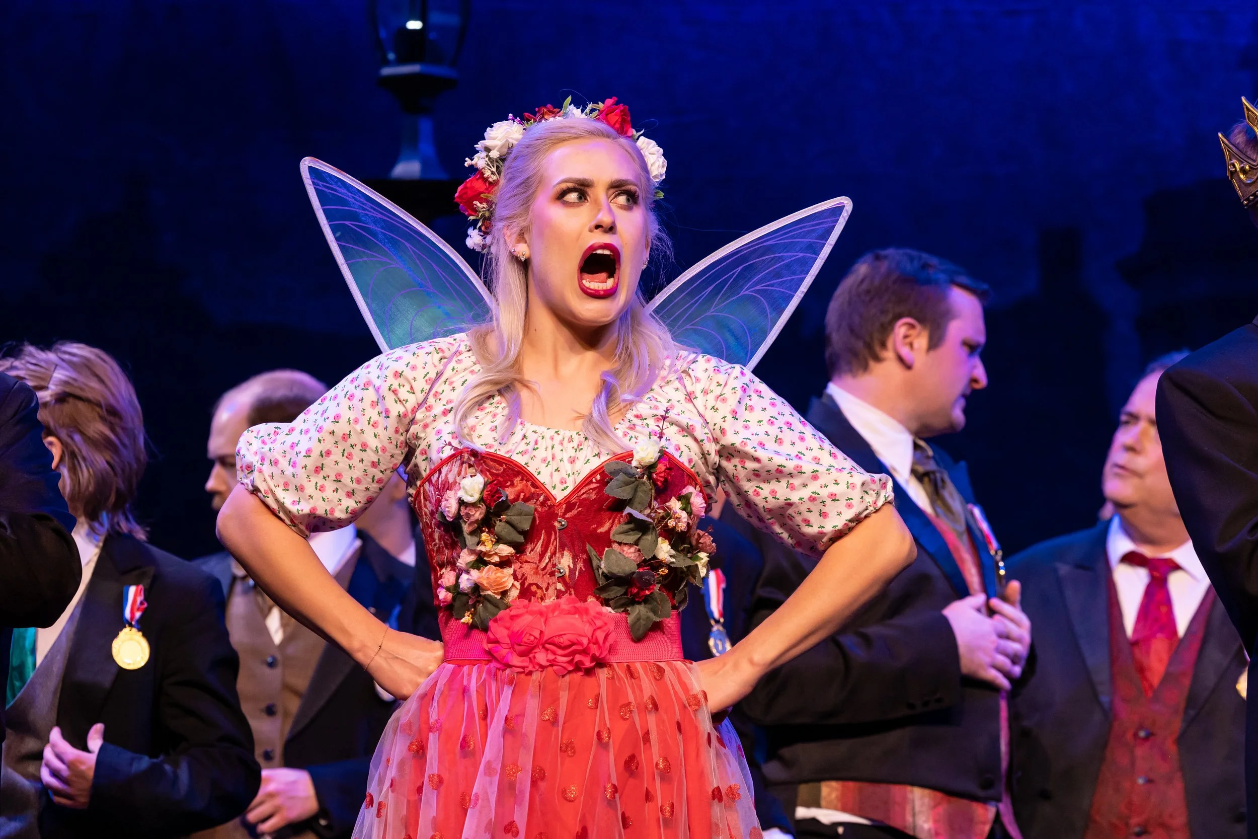 A woman dressed as a fairy with blonde hair, wings, and a floral crown, appears angry or surprised at an event with people in formal attire in the background.