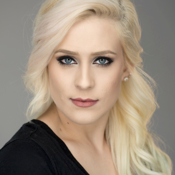 Close-up portrait of a woman with long blonde hair, blue eyes, makeup, and wearing pearl earrings against a gray background.