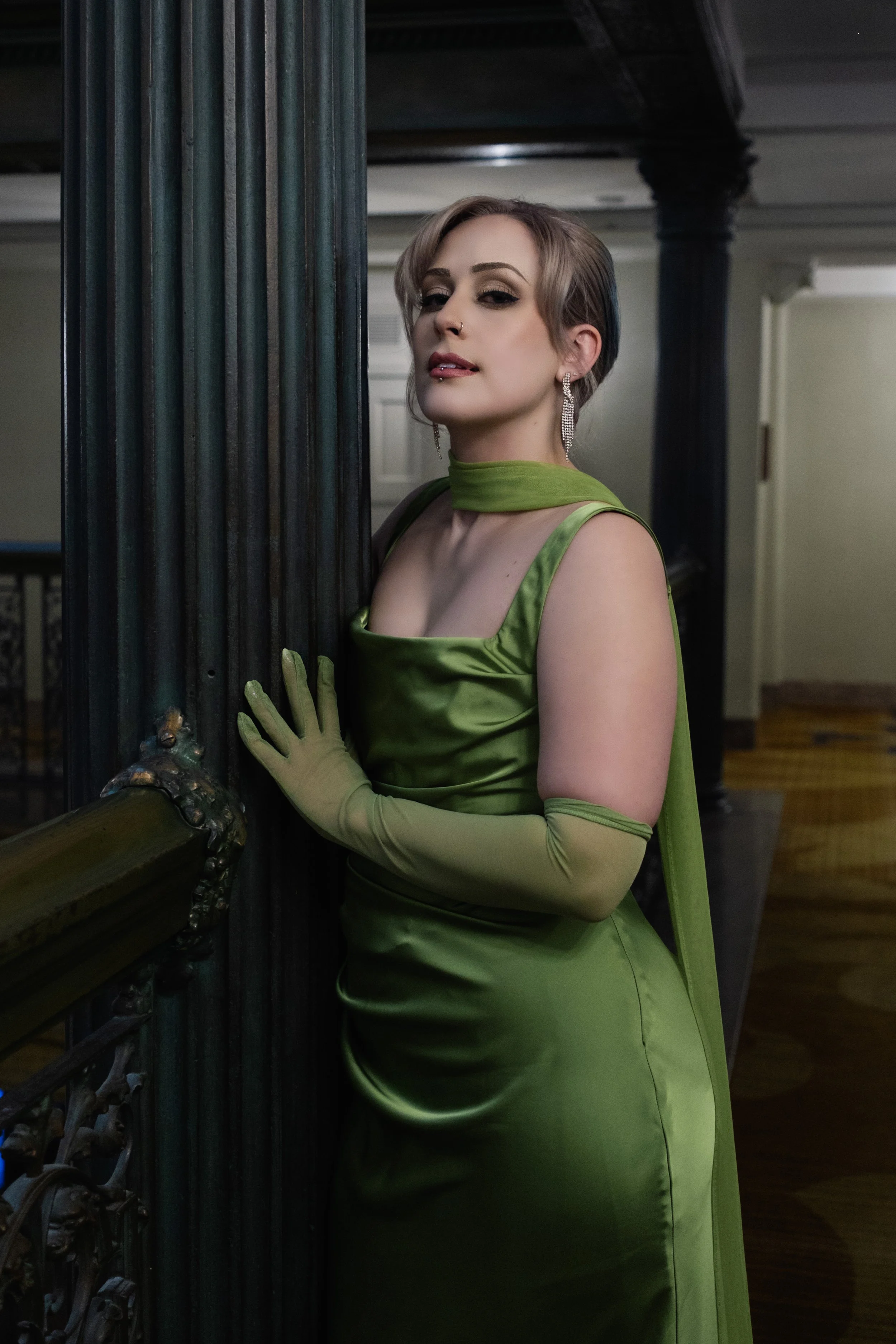 A woman with short hair and makeup in a green satin dress with matching gloves, standing indoors beside a black ornate column.