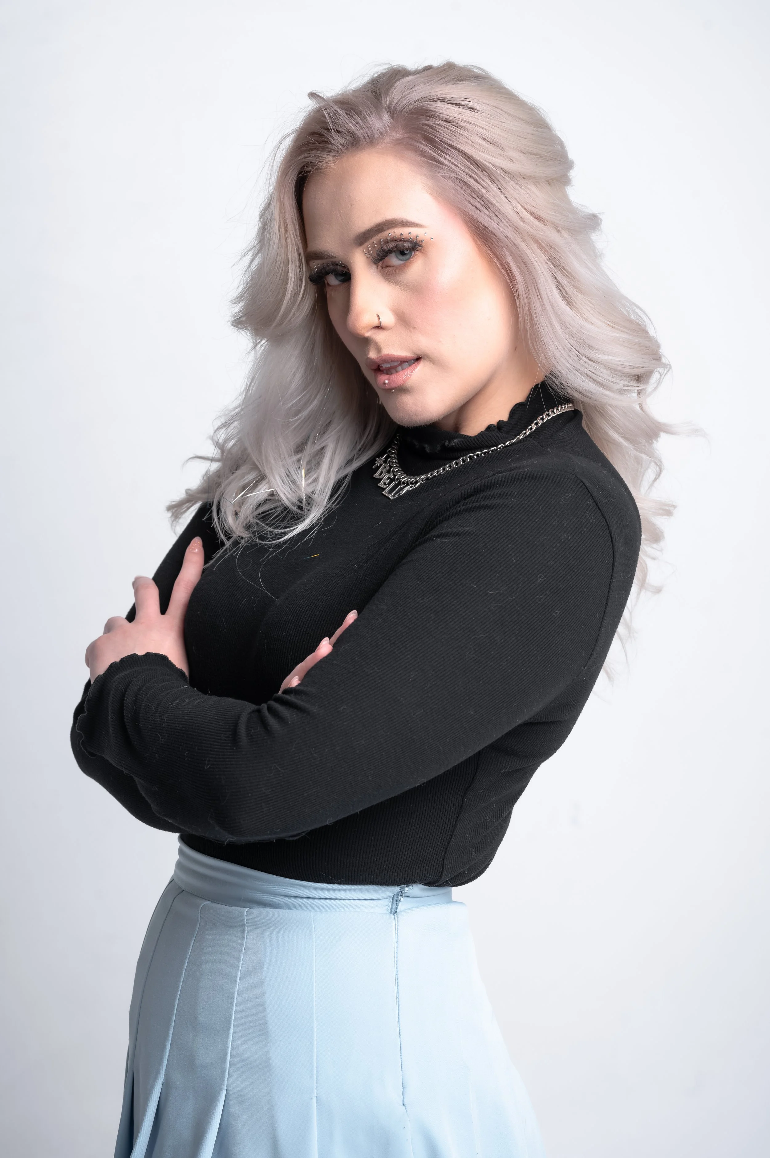 A young woman with light purple hair and makeup featuring rhinestones around her eyes, wearing a black top with chain necklace, crossed arms, and a high-waisted light gray skirt against a plain white background.
