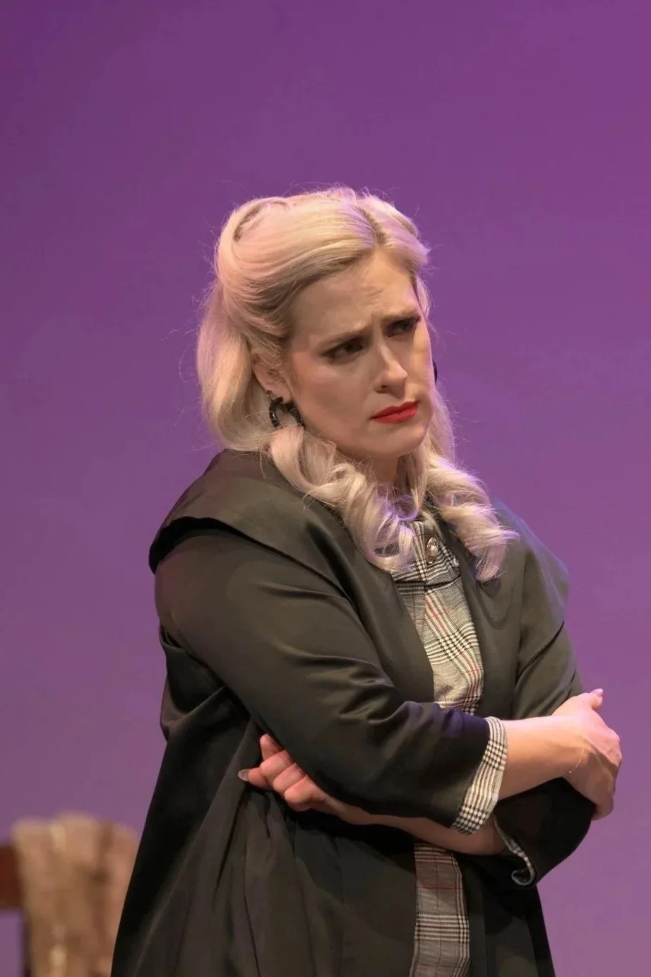 A woman with platinum blonde hair styled in loose curls, wearing a black jacket over a plaid shirt, standing with her arms crossed and a concerned or skeptical expression against a purple background.