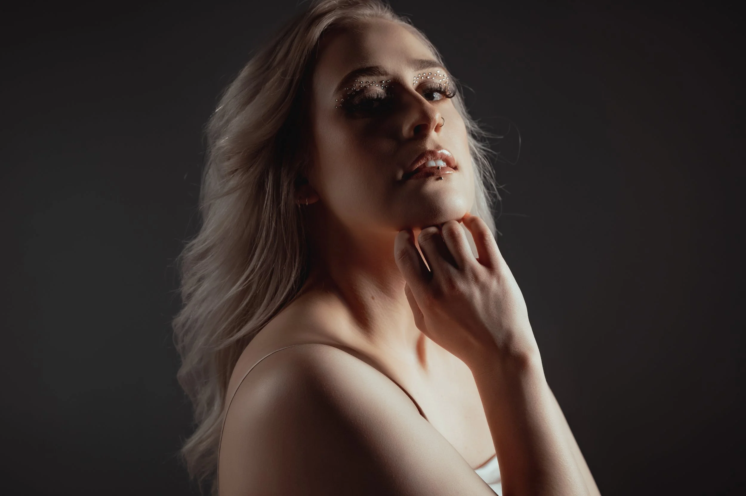 A woman with platinum blonde hair styled in loose waves, wearing makeup embellished with rhinestones above her eyebrows, with a contemplative expression and her hand gently touching her chin, against a dark background.