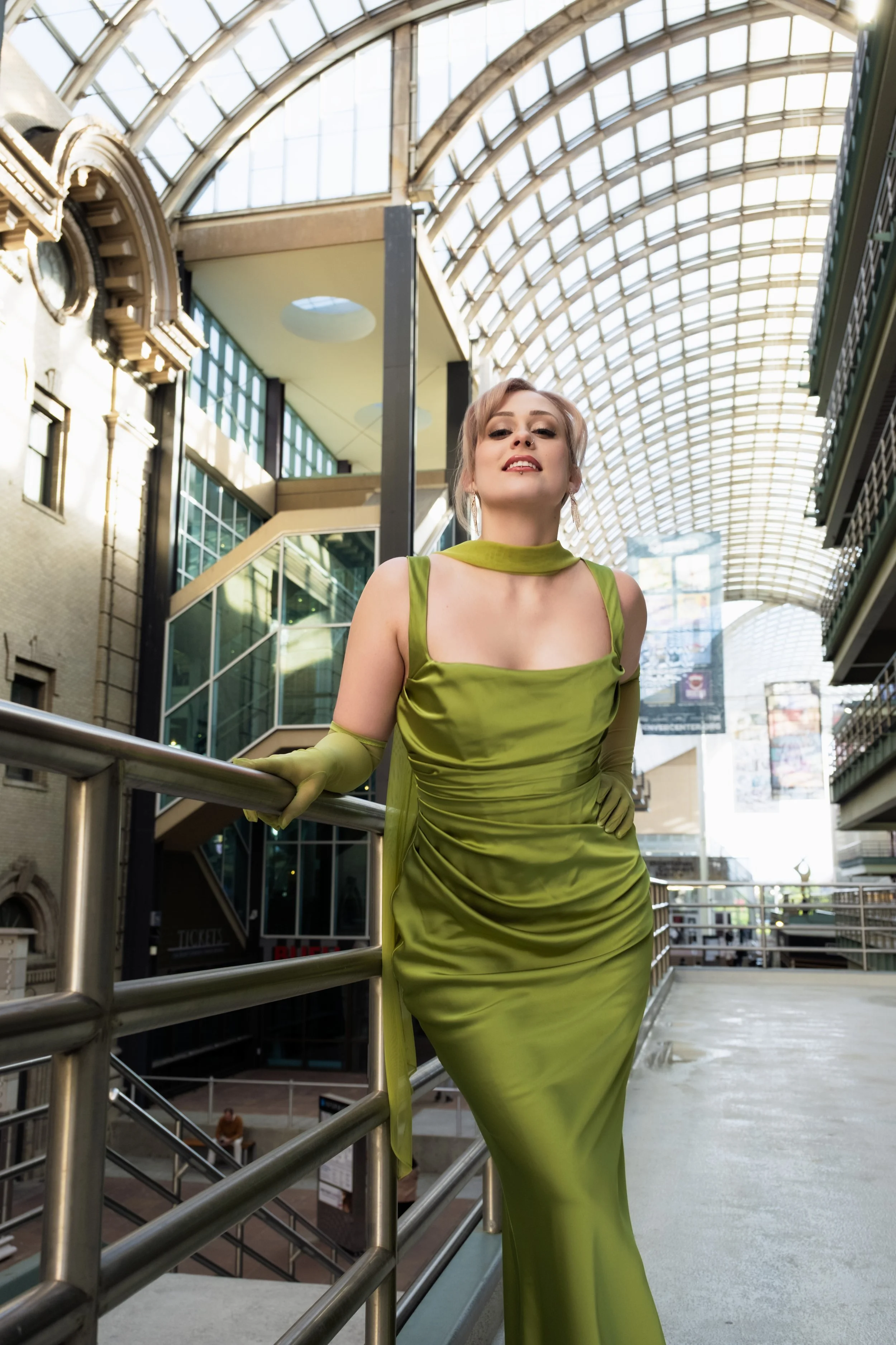 Woman in a shiny green dress and matching gloves posing in an indoor shopping gallery with a glass and steel arched ceiling.