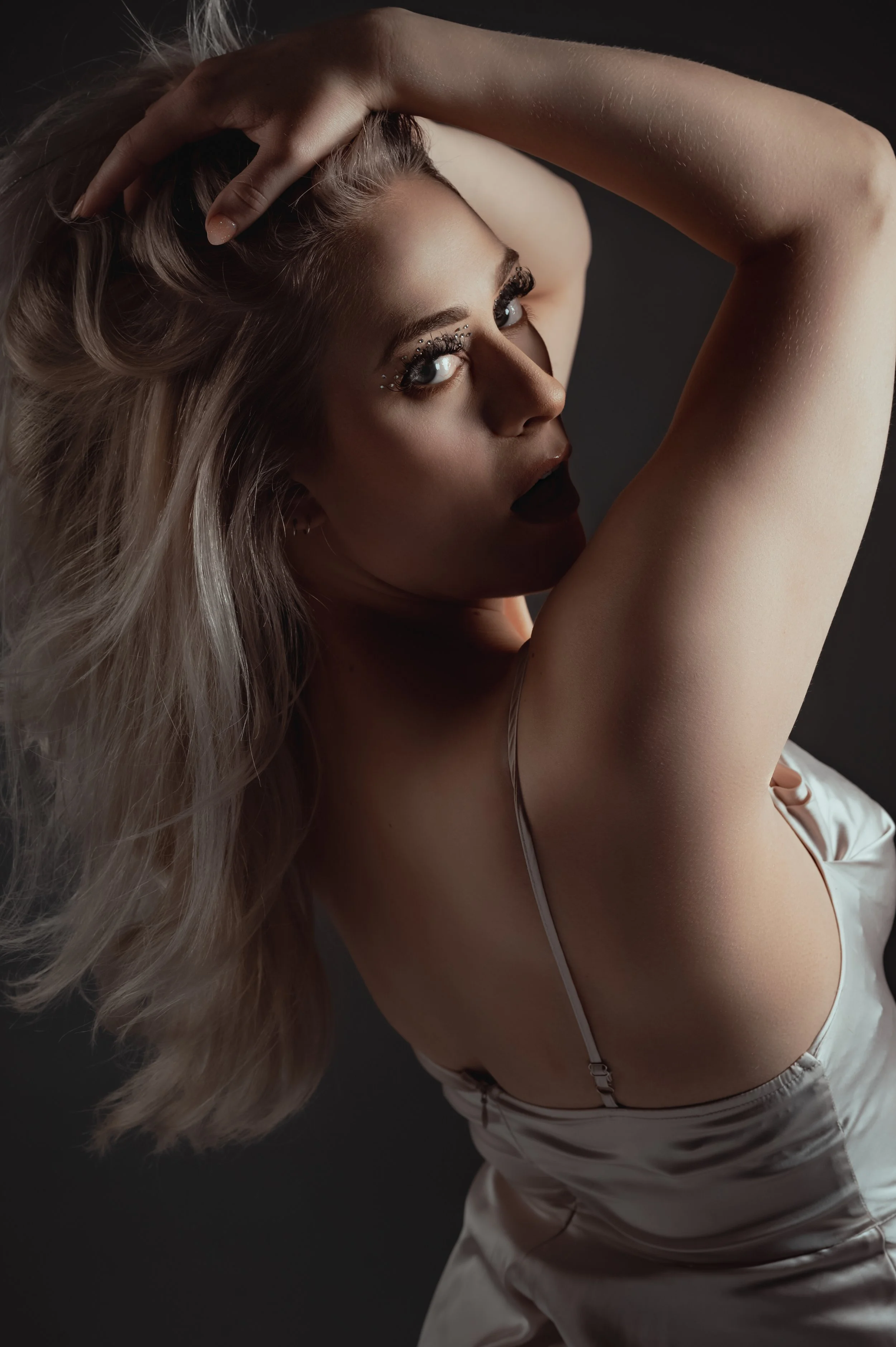A woman with long blond hair posing with her arm above her head, wearing makeup with decorative rhinestones near her eyes, in a sleeveless satin top, against a dark background.