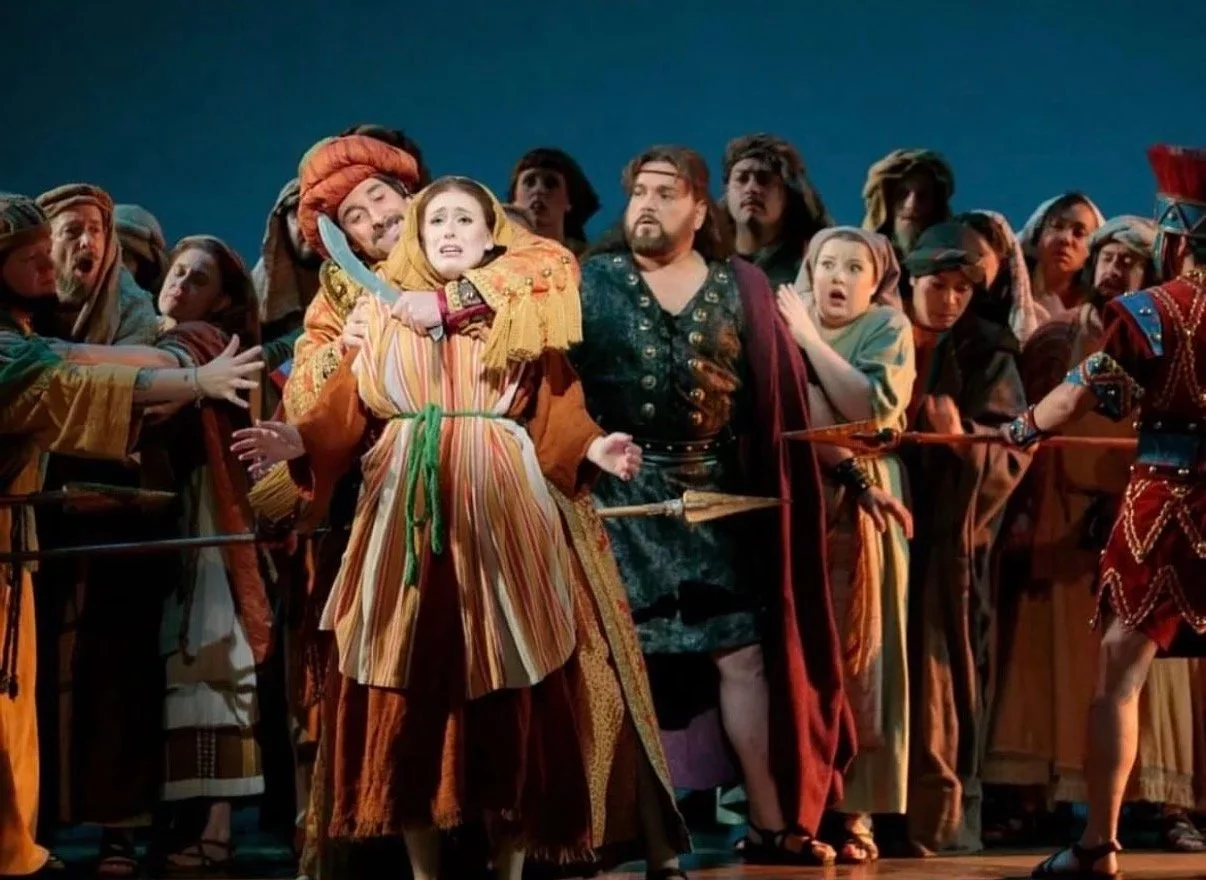 Scene from a theatrical performance with actors in costumes, depicting a dramatic moment with one character holding a sword and others showing expressions of alarm and concern.