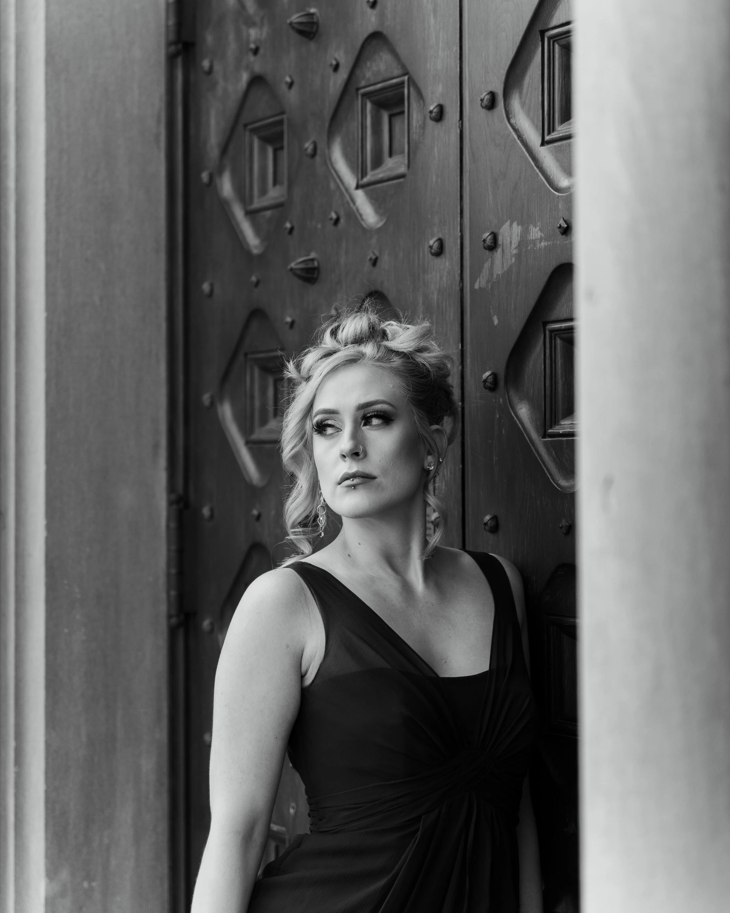 Black and white photo of a woman with blonde hair in an updo, wearing a dark dress, looking to her left, standing in front of a large wooden door with decorative patterns.