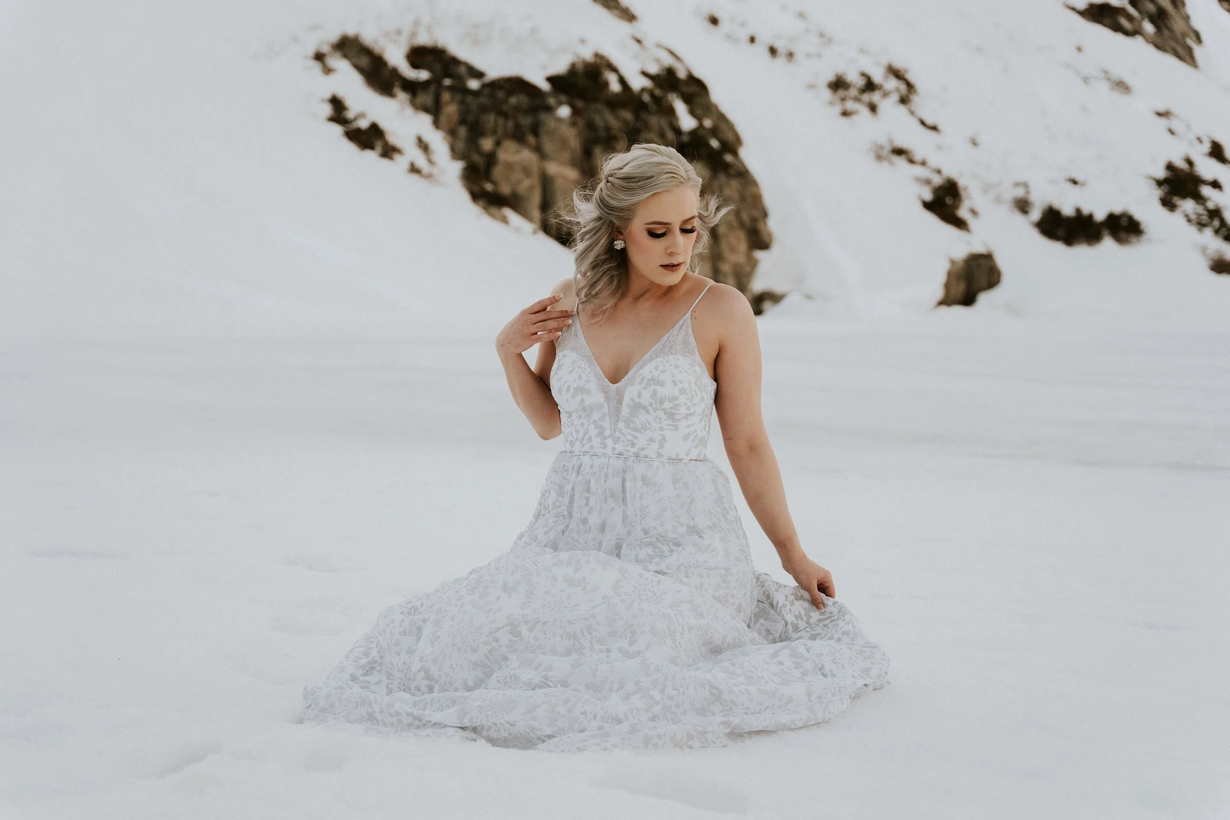 A woman with long, wavy gray hair in a loose updo, wearing a white lace wedding dress with spaghetti straps, standing in a snowy landscape with snow-covered rocks in the background.