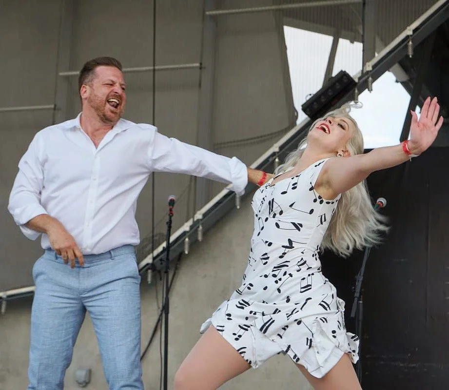 A man pulling a woman by her arm on a stage, both laughing and enjoying themselves, with microphones in the background.
