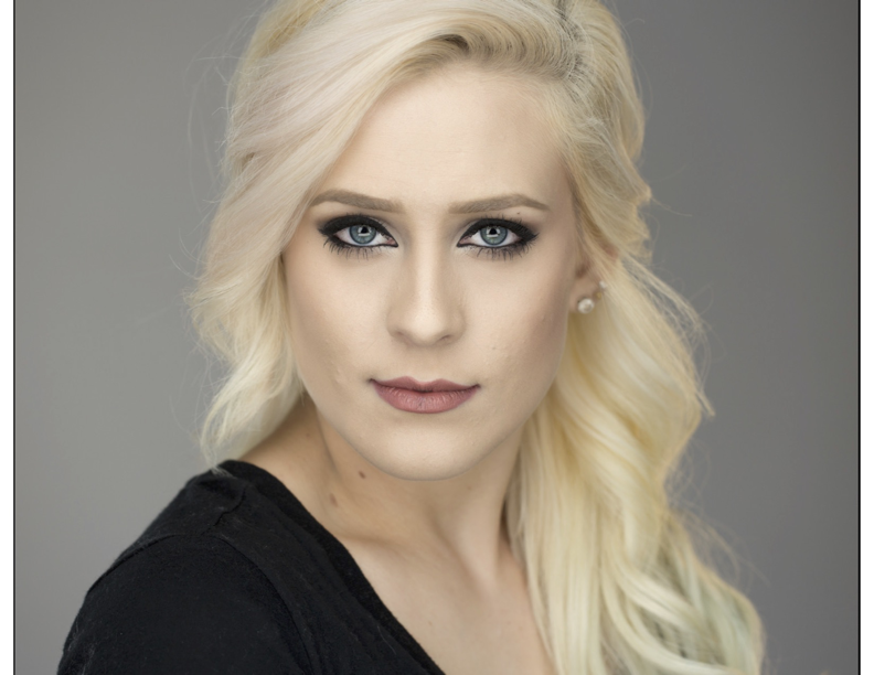 Close-up portrait of a blonde woman with blue eyes, makeup, and a black top against a gray background.