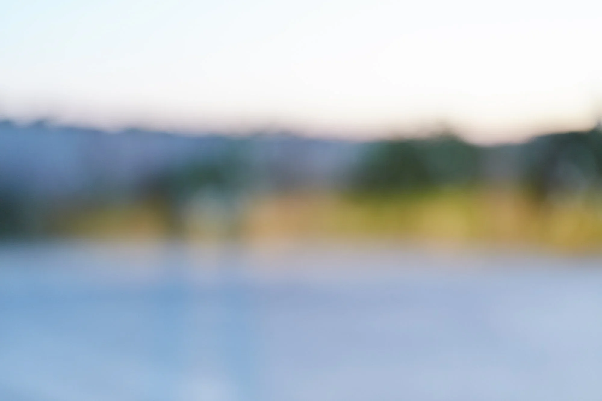 Blurred landscape scene with a body of water, green trees, hills, and a clear sky.