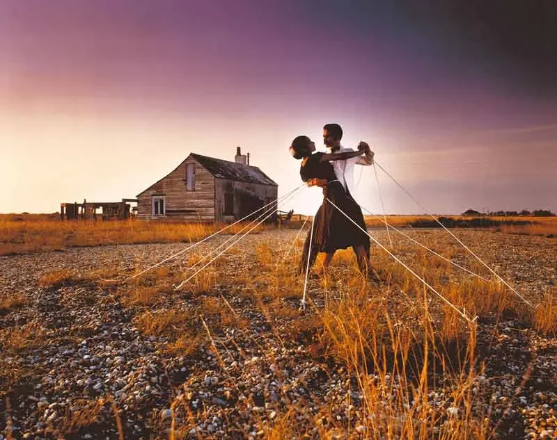 A scenery with two women in vintage clothing with parachute strings attached, standing in a dry, grassy field near an old, weathered house at sunset.