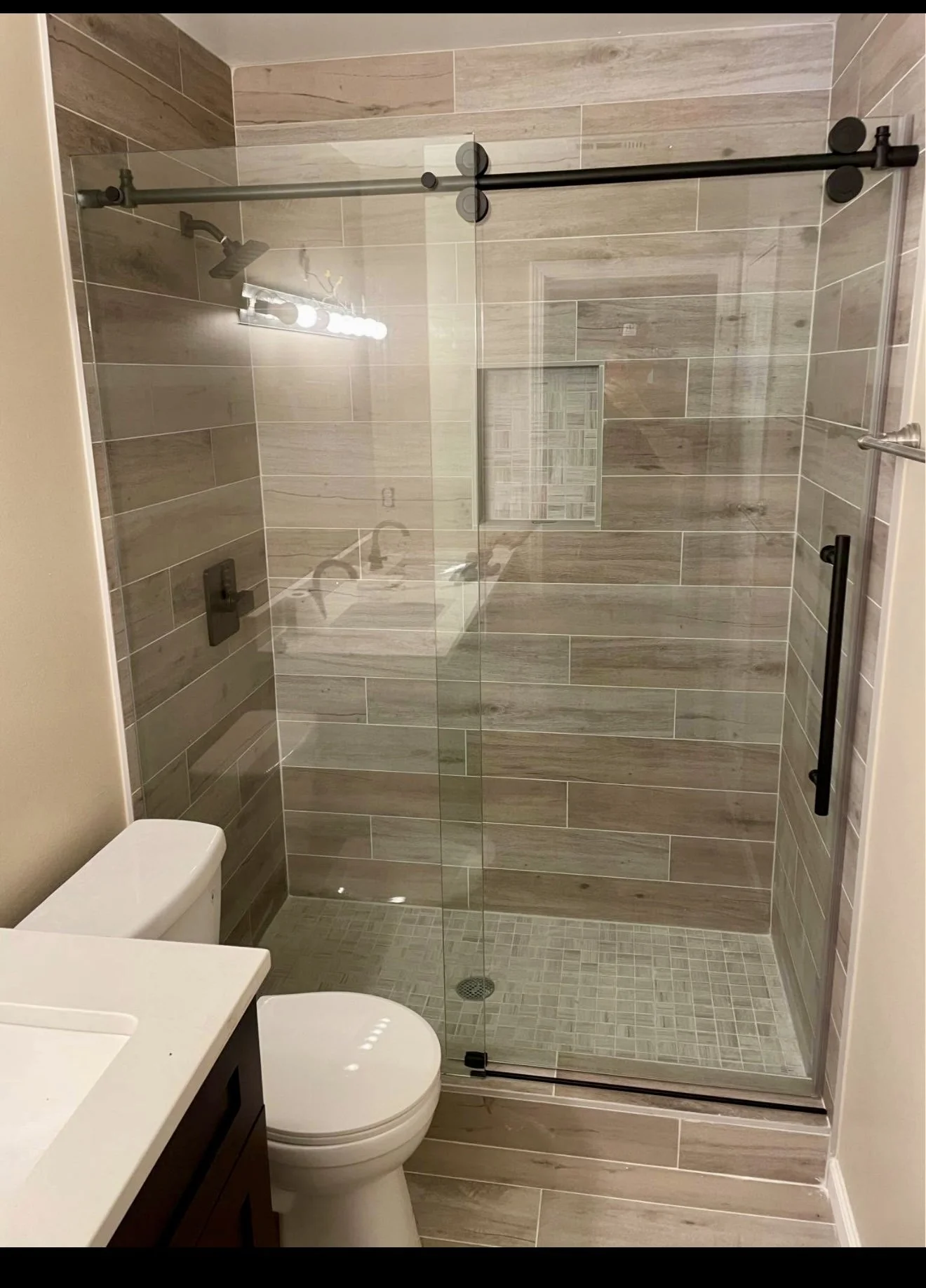 Walk-in shower finished with wood-look wall tile, mosaic flooring, a built-in niche, and sliding glass enclosure.