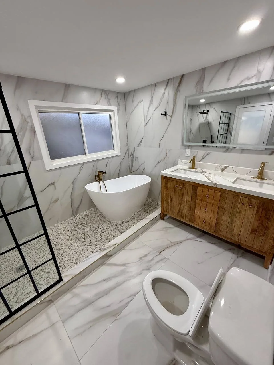 Complete bathroom remodel featuring marble-look surfaces, freestanding soaking tub, double vanity, and modern black and brass accents.