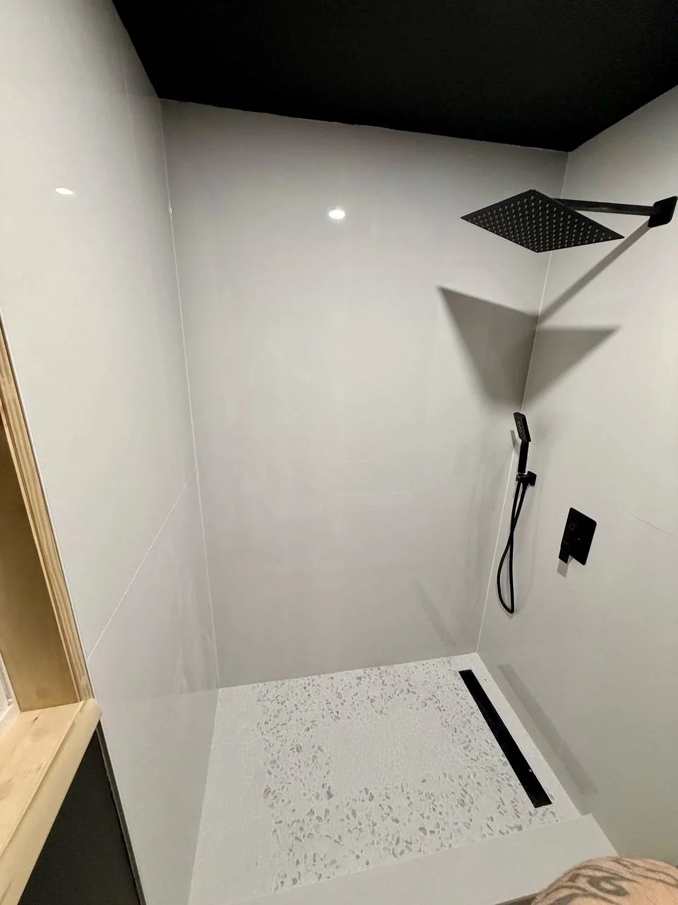 Large-format white wall tile, pebble-style shower floor, linear drain, and matte black rainfall fixtures create a minimalist shower.