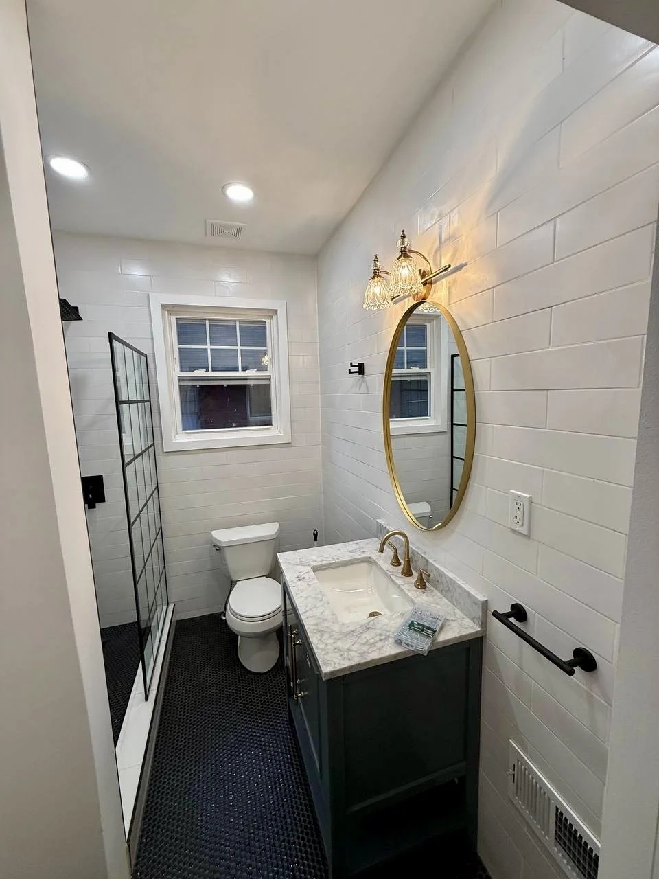 Finished bathroom featuring white tiled walls, black mosaic flooring, dark vanity, marble countertop, and warm brass accents.