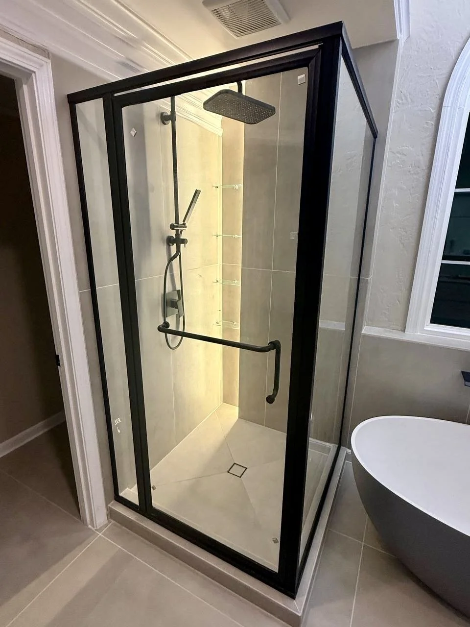 Large-format neutral tile, black-framed glass, rainfall showerhead, and integrated shelving complete this modern shower.