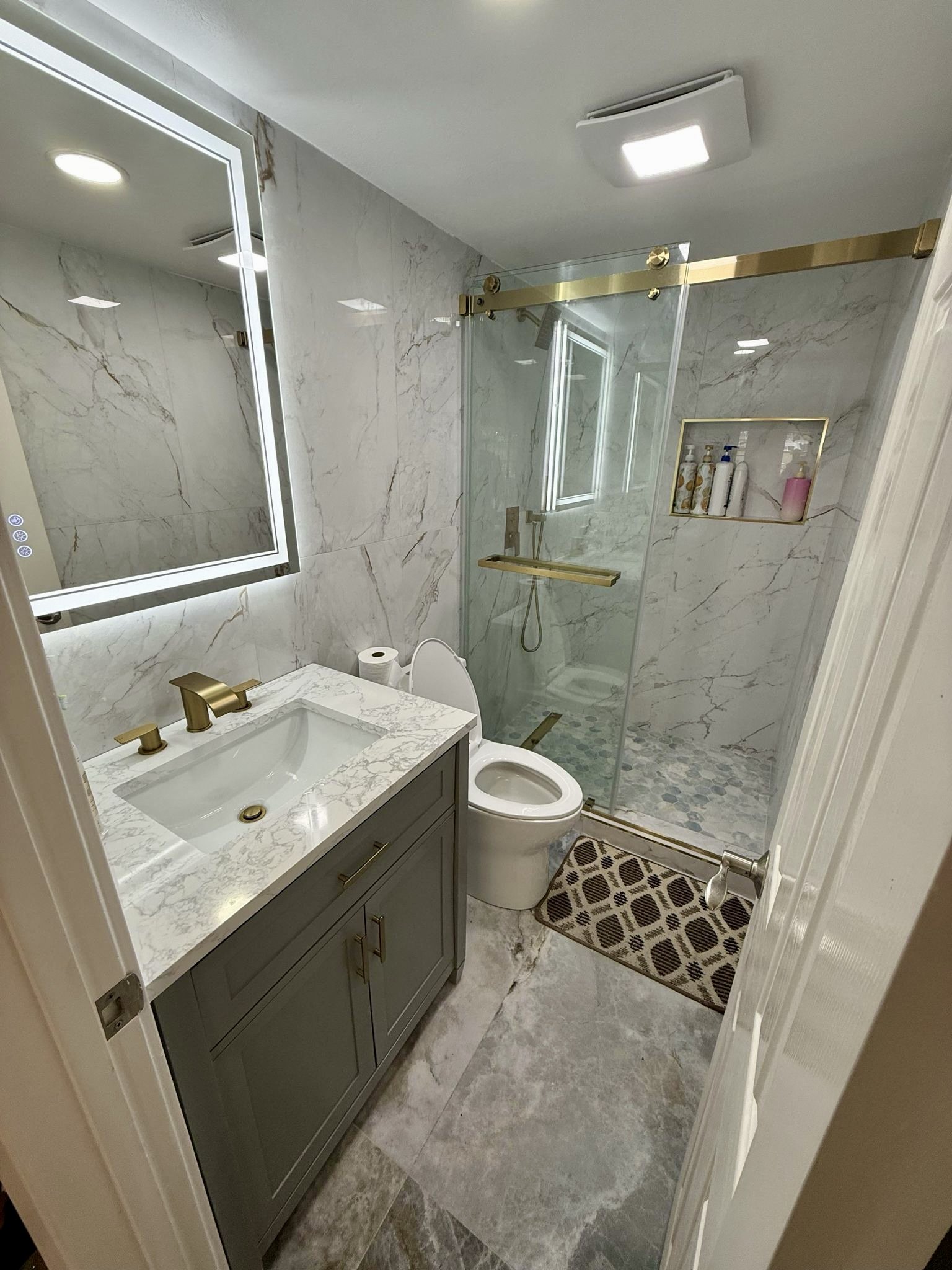 Complete bathroom renovation featuring marble-look tile, a glass-enclosed walk-in shower, built-in niche, gray vanity, illuminated mirror, and coordinated brushed-gold fixtures.