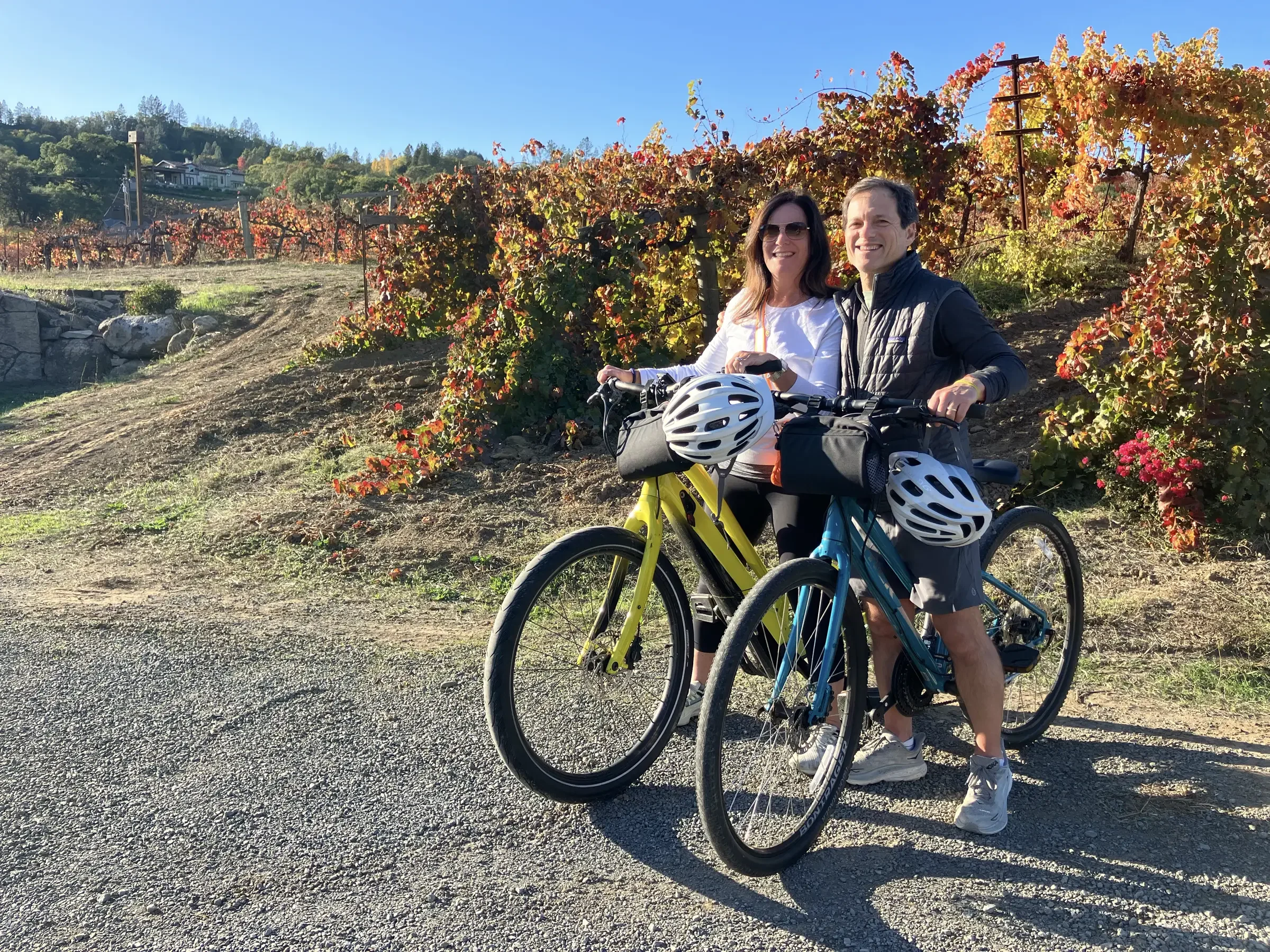 A man and a woman smiling while standing next to their bicycles on a dirt path near vineyard rows with autumn leaves on the vines, under a clear blue sky.