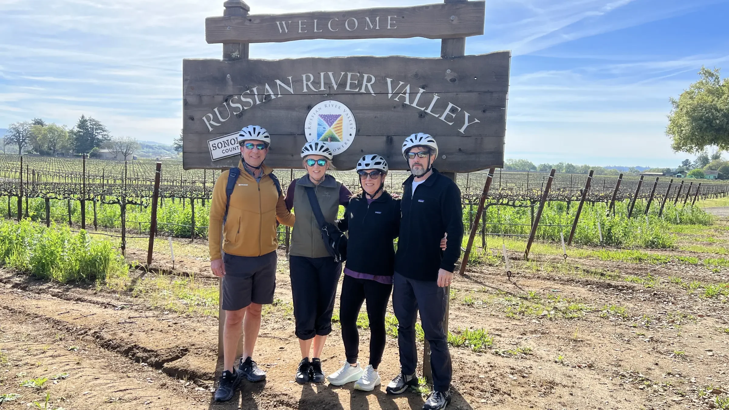 Four people wearing helmets and sunglasses standing in front of a large wooden sign that reads "Russian River Valley" at a vineyard.