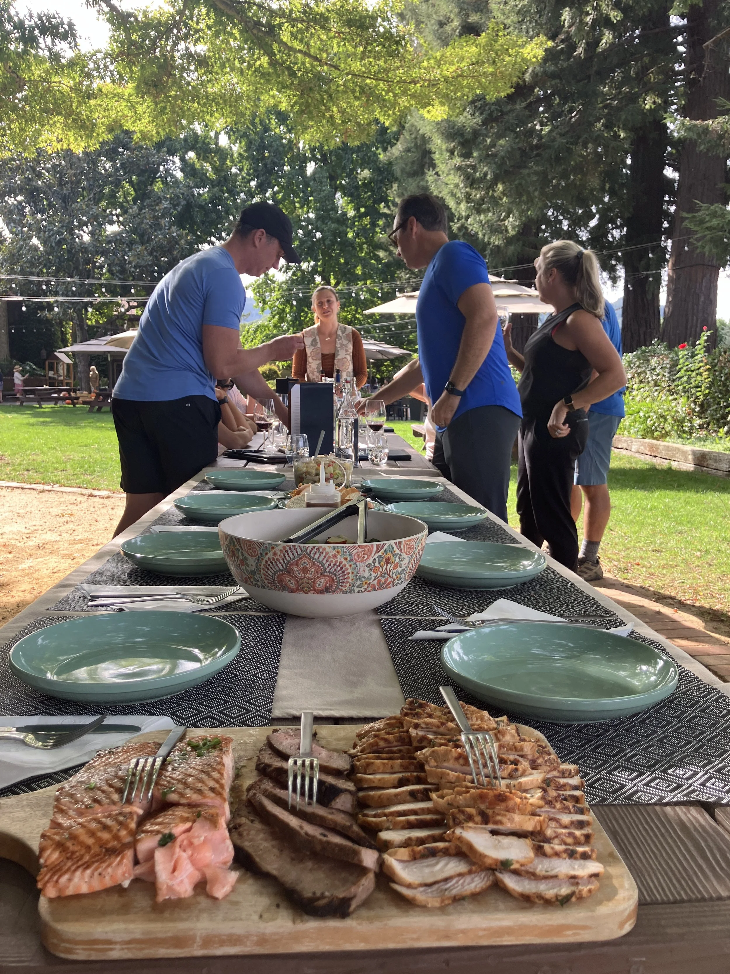 A long outdoor dining table set with empty green plates, cutlery, and a large platter of sliced grilled meats, with people gathered around under trees in a park.
