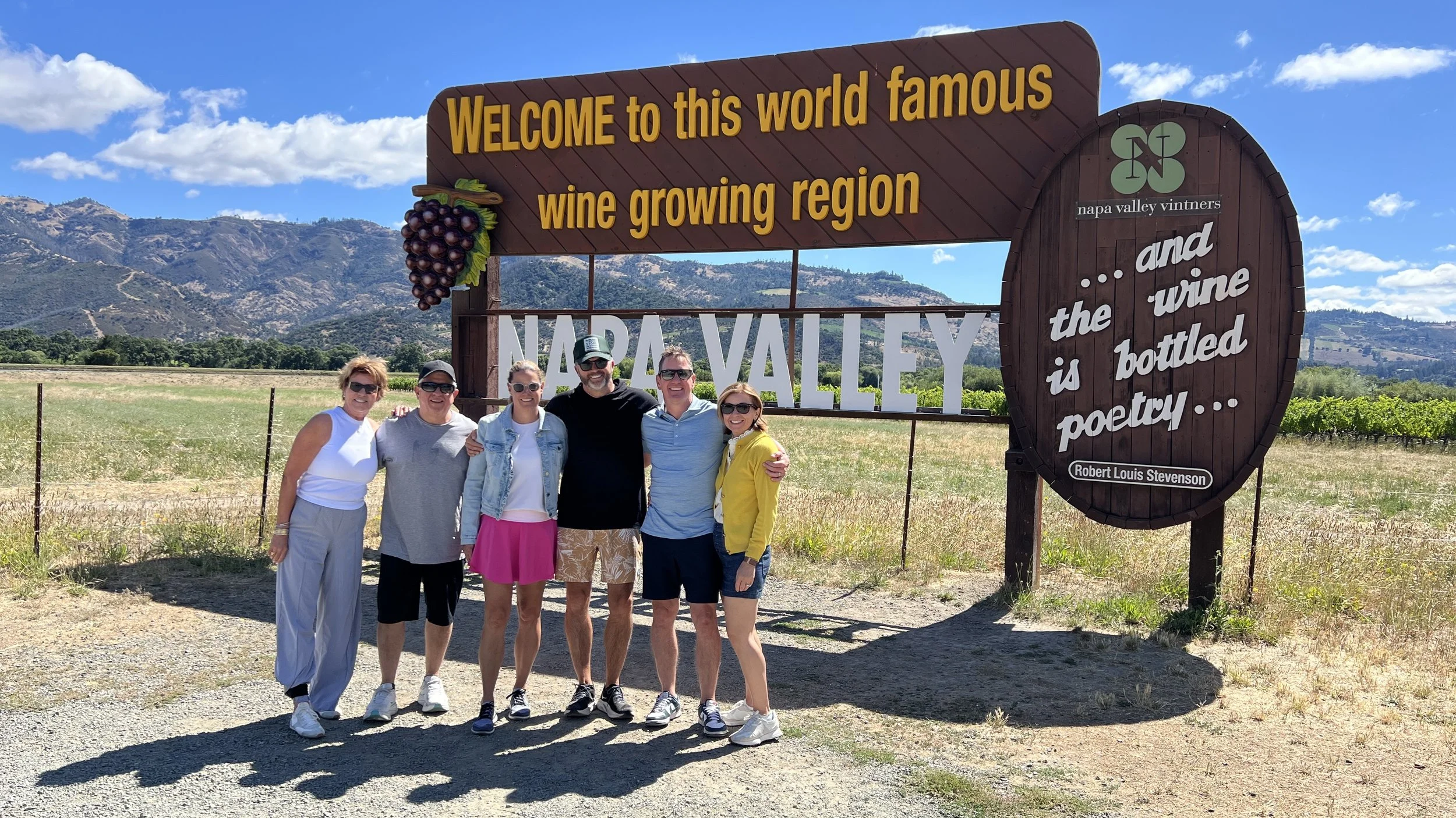 Group of six people standing in front of a large welcome sign at Napa Valley Winery, with hills and blue sky in the background.