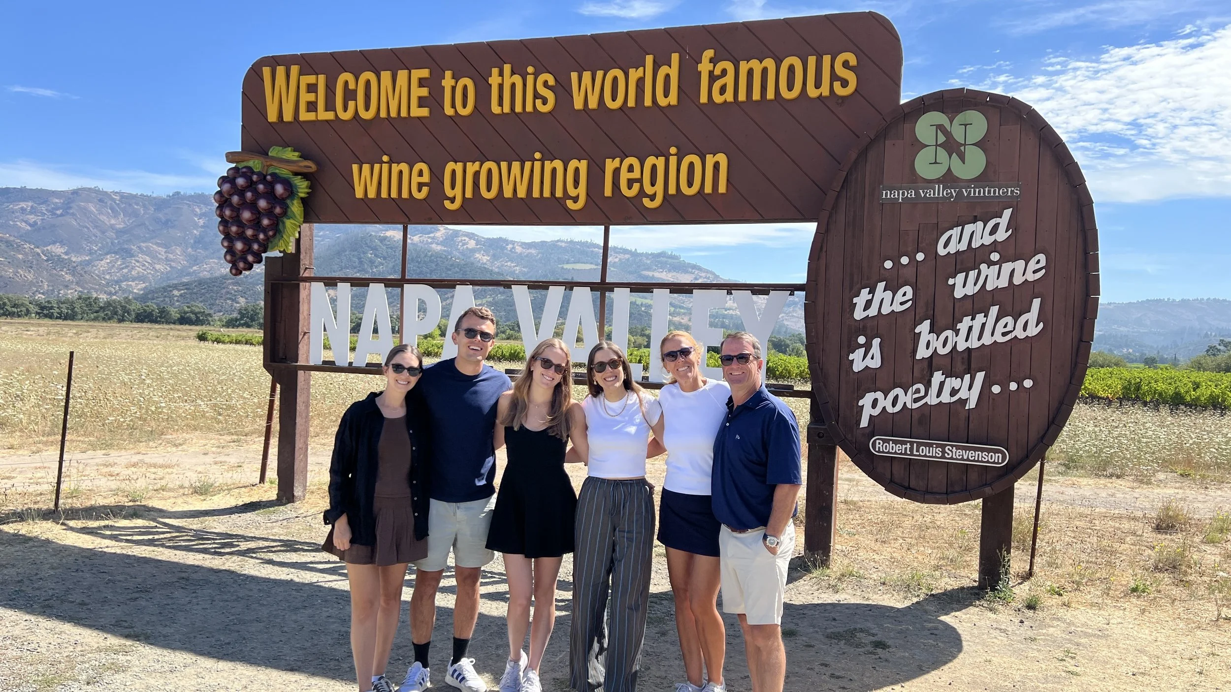 Group of six smiling people standing in front of a wooden sign that says, 'Welcome to this world famous wine growing region Napa Valley and the wine is bottled poetry' with mountains in the background.