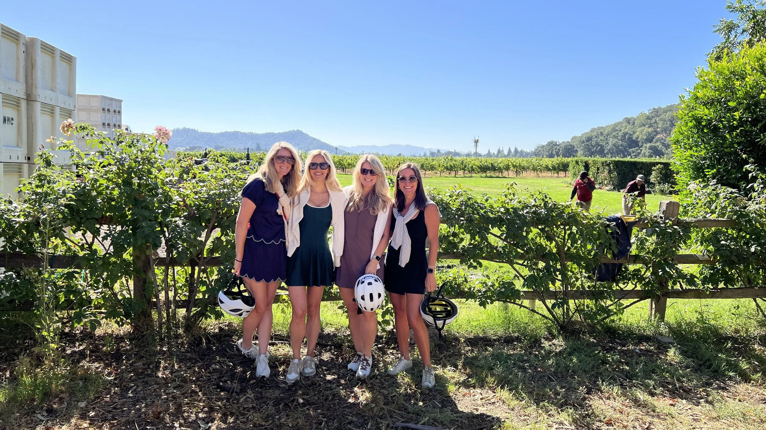 Four women standing outdoors in front of a vineyard, smiling, holding bicycle helmets, dressed casually, with greenery and distant hills in the background.