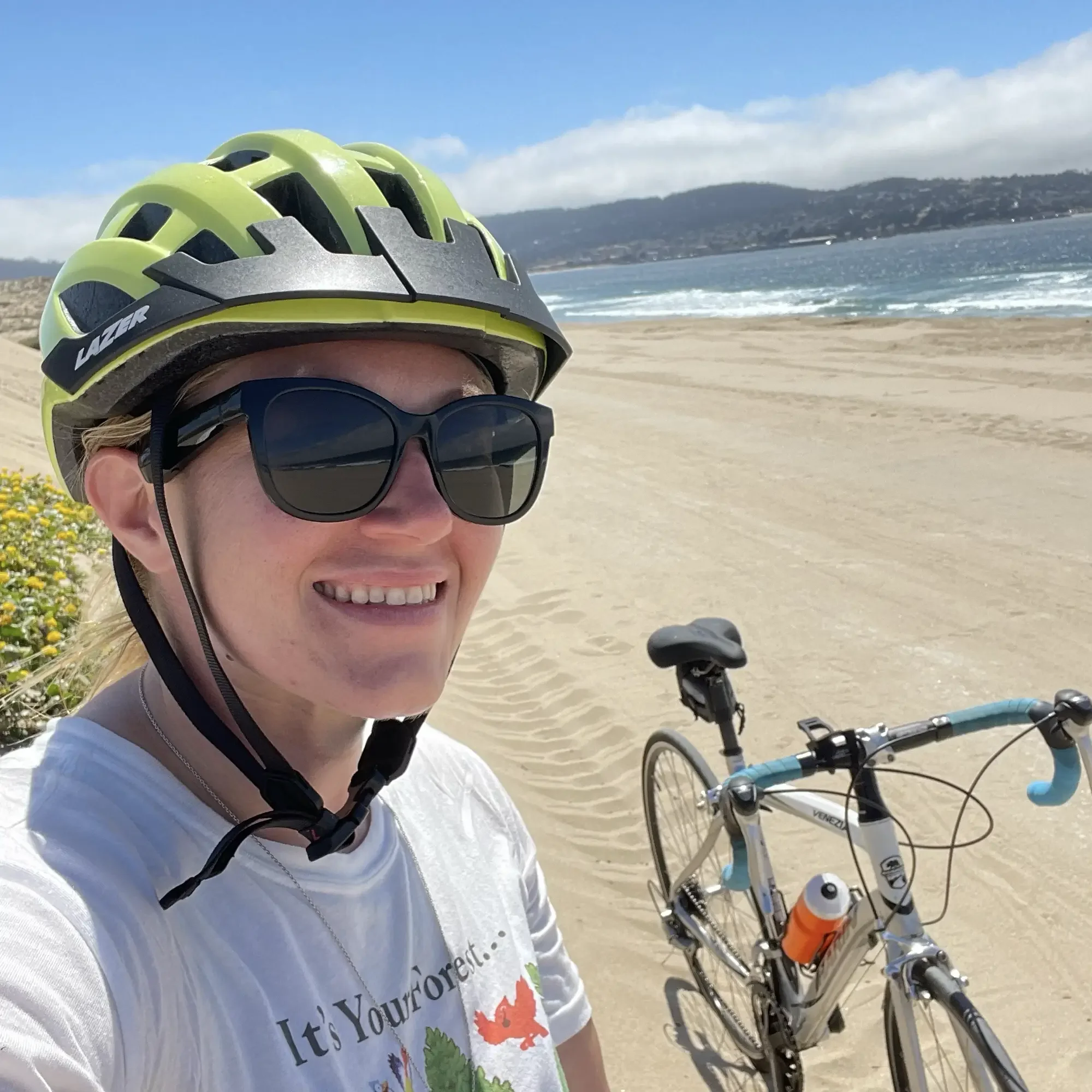 Person wearing a green bike helmet and sunglasses taking a selfie at the beach with a bicycle, ocean waves, and a distant hill in the background.
