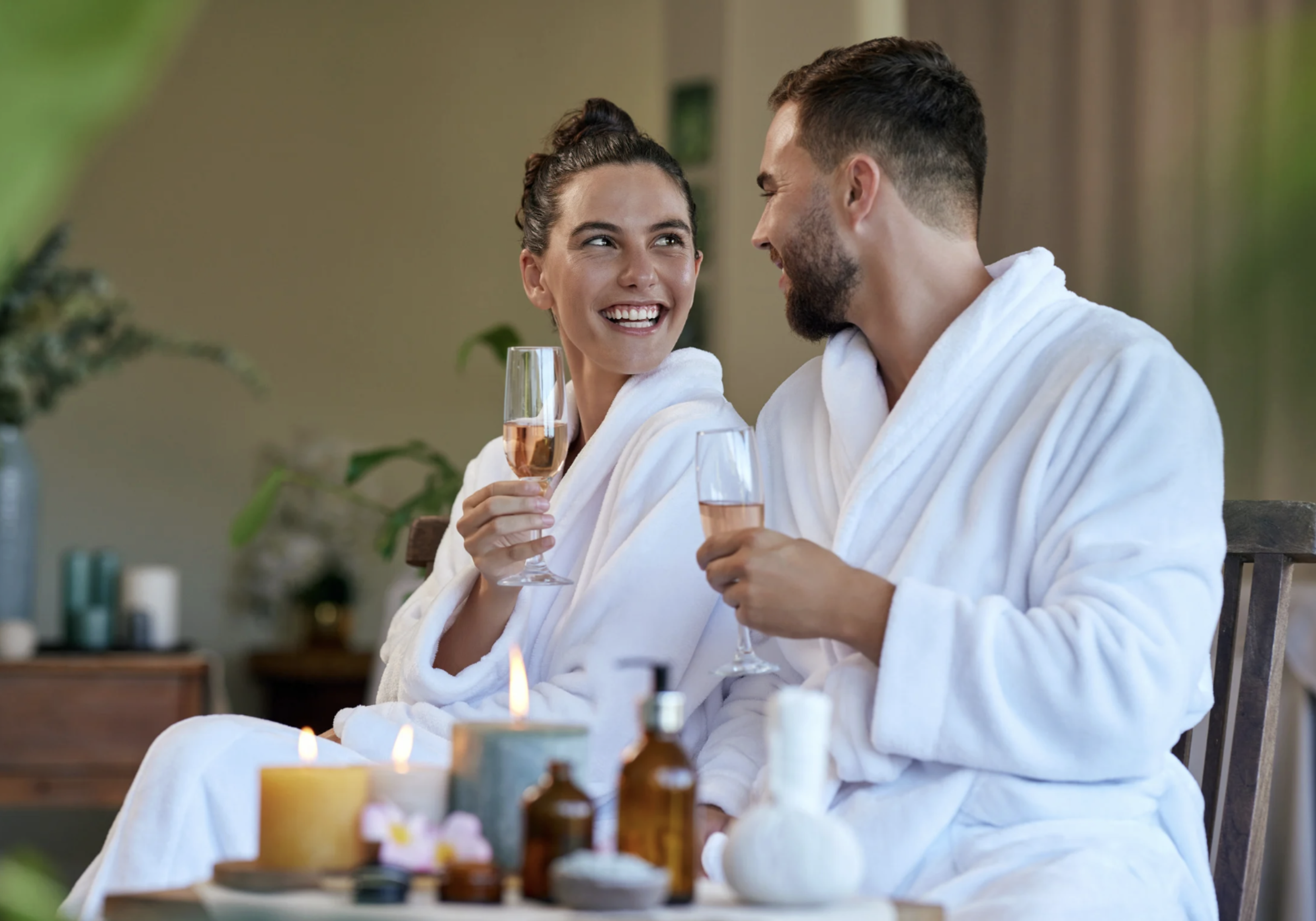 A happy couple in bathrobes celebrating with champagne at a spa or wellness center with candles and skincare products on the table.