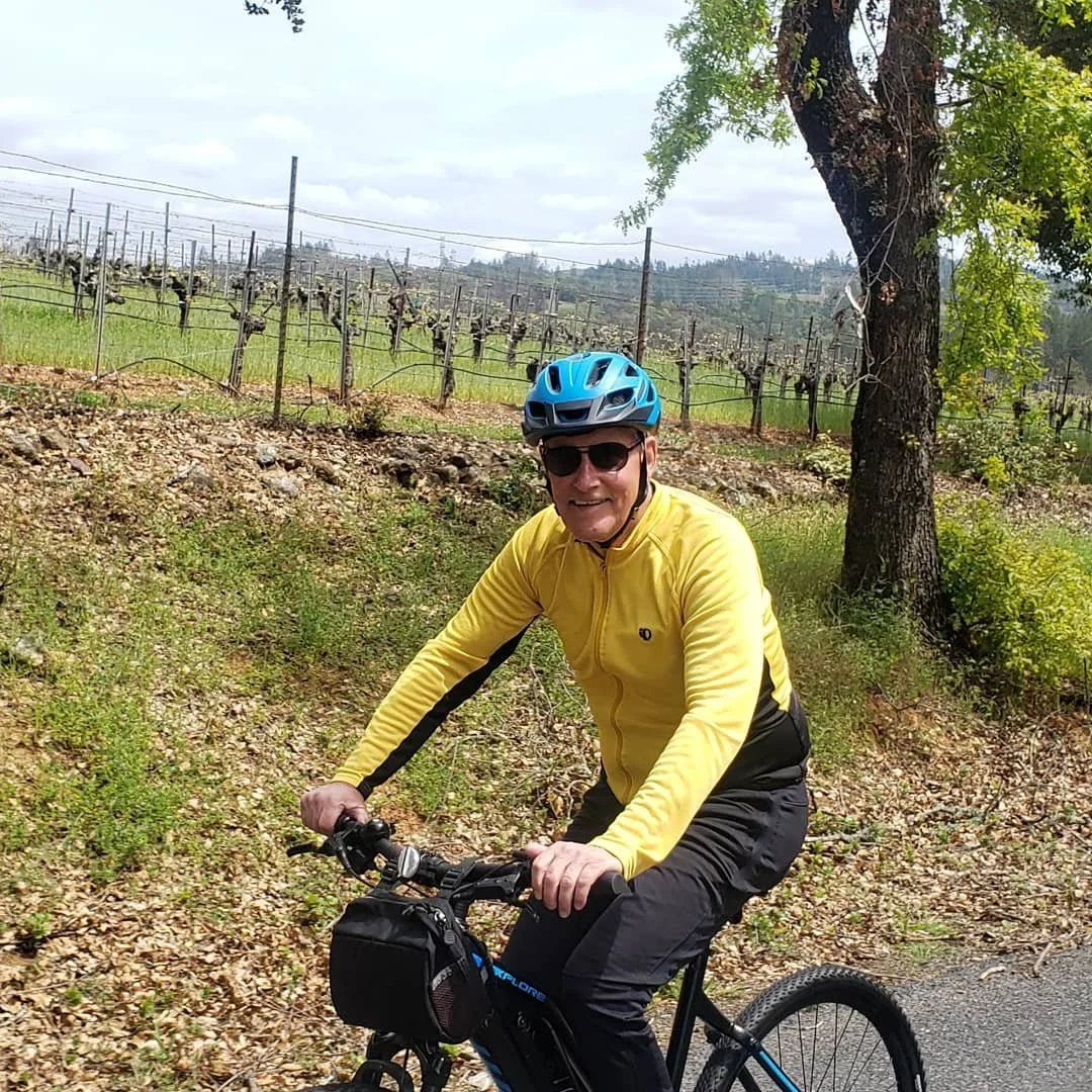 Dave, the owner of Adventures in Cycling,  riding a bicycle along a trail next to a vineyard with leafless grapevines and a large tree nearby.