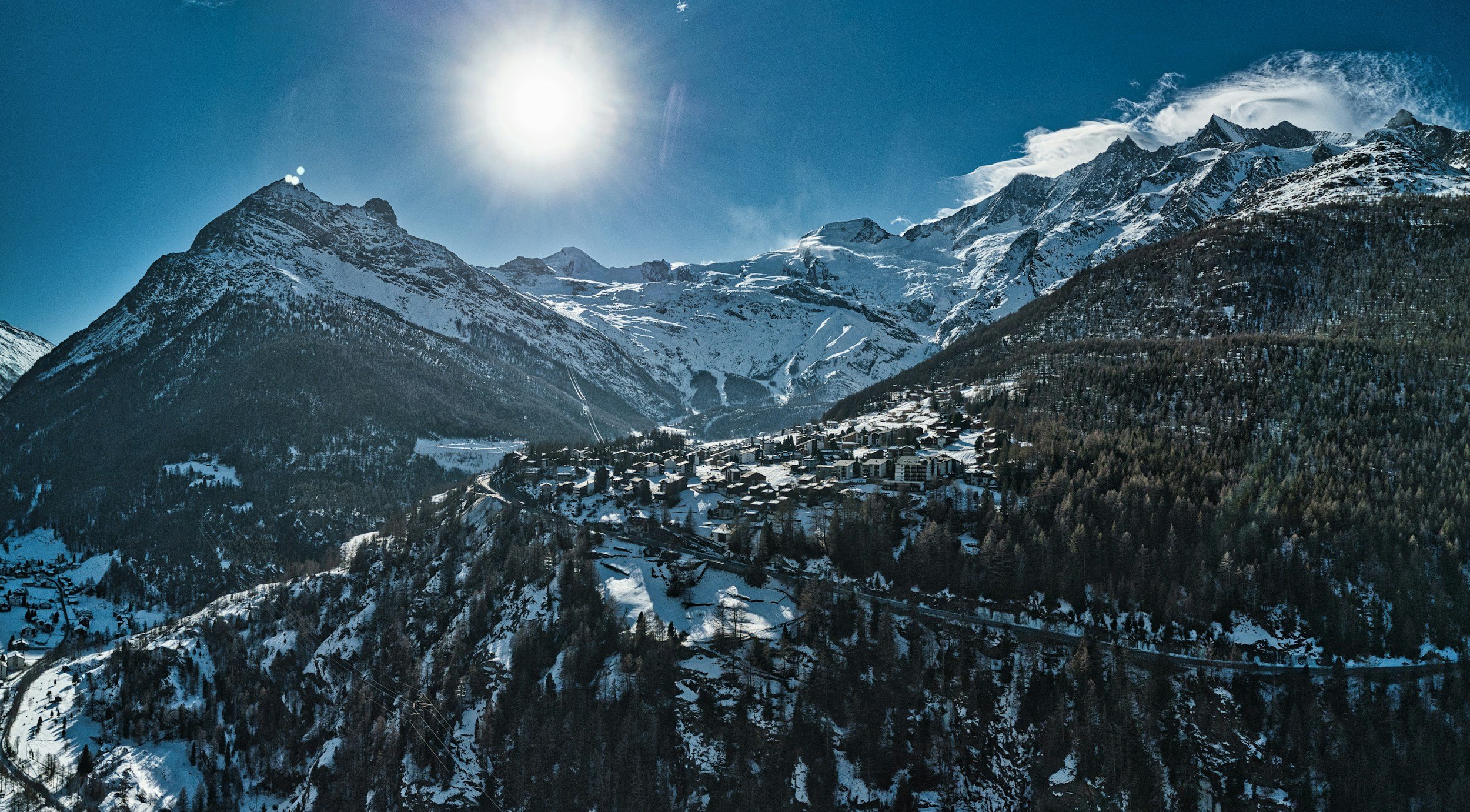 Snow-covered mountain landscape with a small village at the base, under a bright sun and clear blue sky.