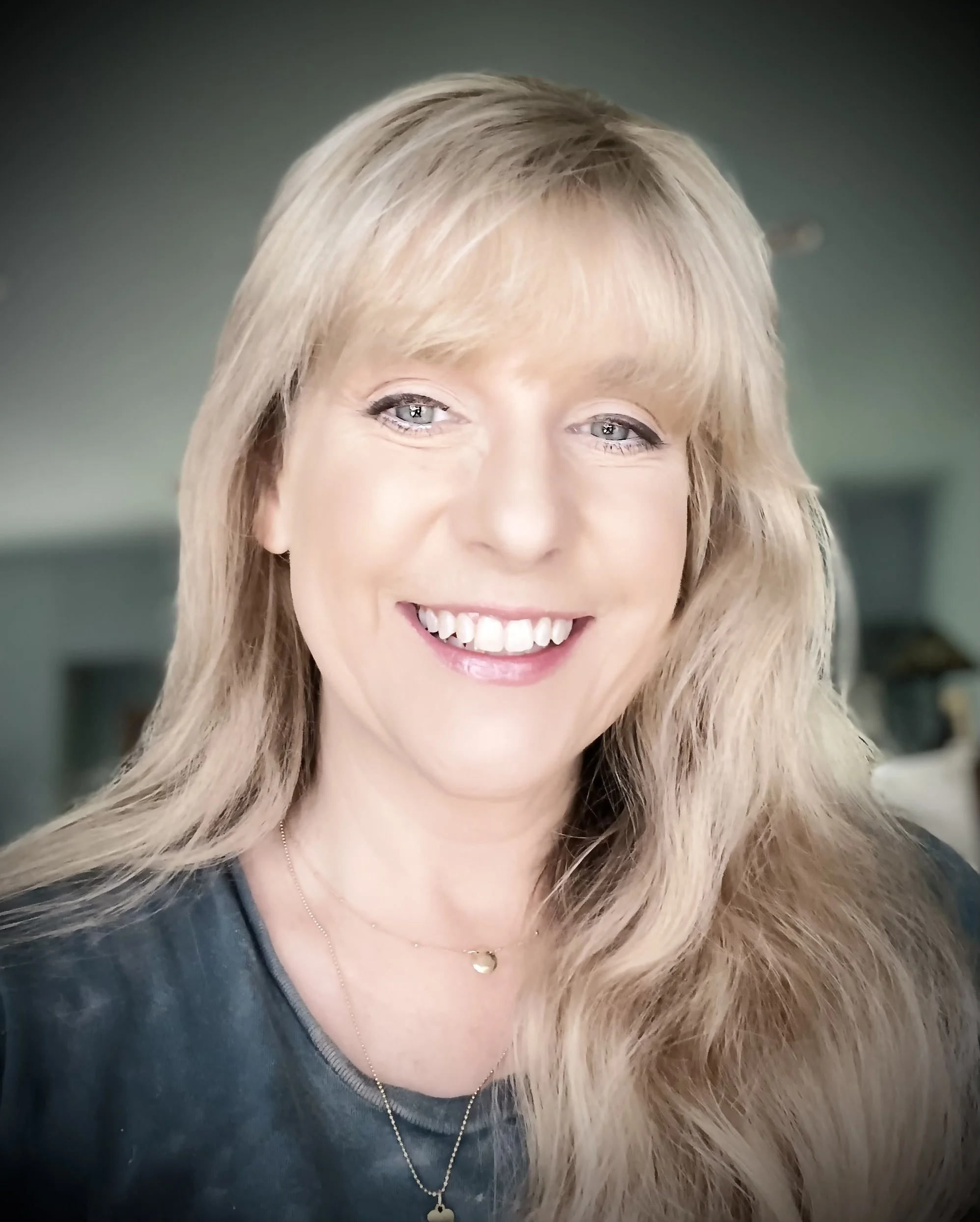 Close-up of a woman with blond hair, blue eyes, smiling, wearing a black top, a heart-shaped pendant necklace, and a delicate chain with a small lock pendant.