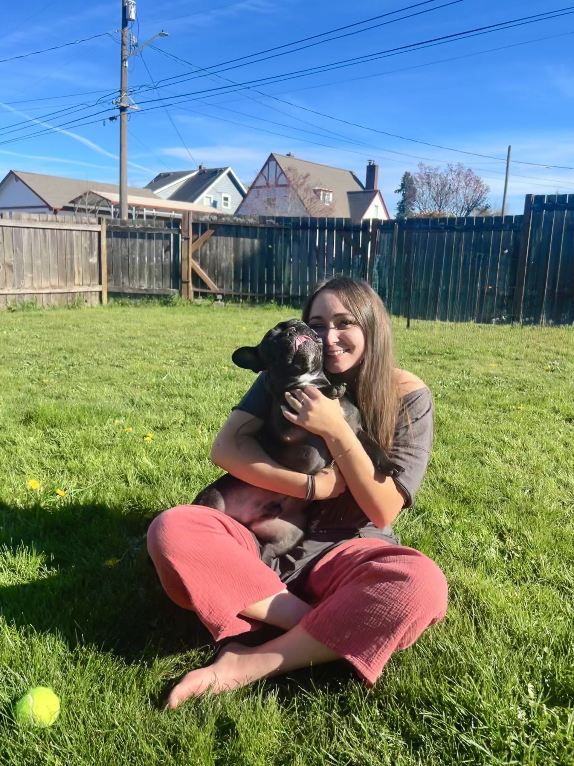 A young woman sitting on grass, smiling and hugging a black French Bulldog with a brindle coat.