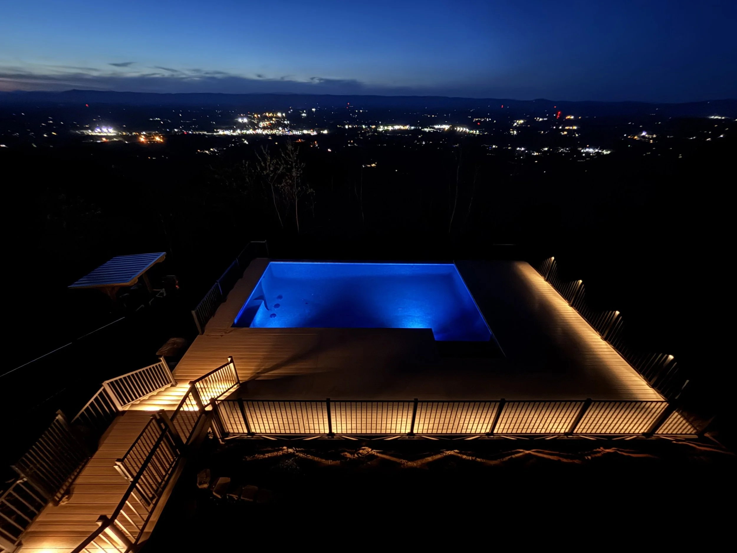 A rooftop hot tub illuminated with blue light overlooking a cityscape at night, with distant mountains under a dark sky.
