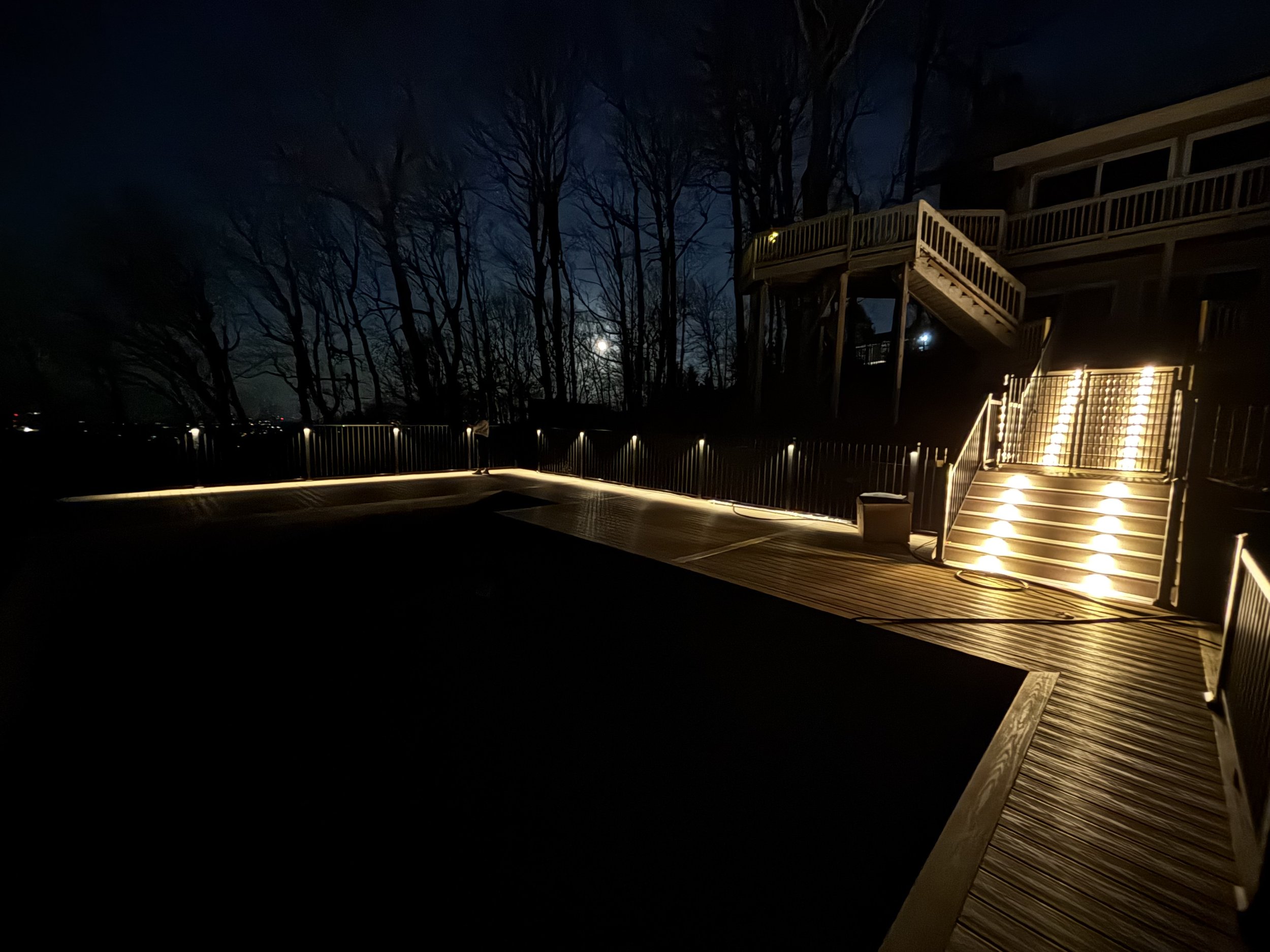 Nighttime outdoor scene showing a wooden deck with stairs and railings illuminated by pathway lights, leafless trees silhouetted against the moonlit sky in the background.