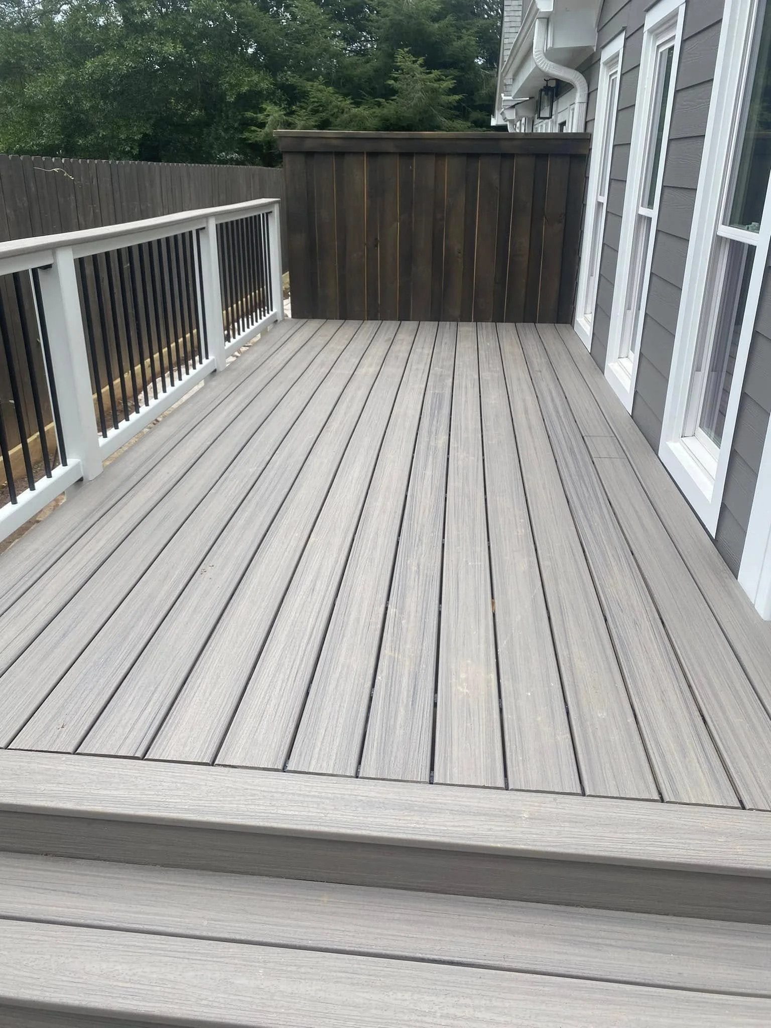 View of a backyard deck made of gray composite wood, with black and white railing on one side and gray siding house with three windows and a door on the other side. A wooden privacy fence is visible in the background with greenery beyond.