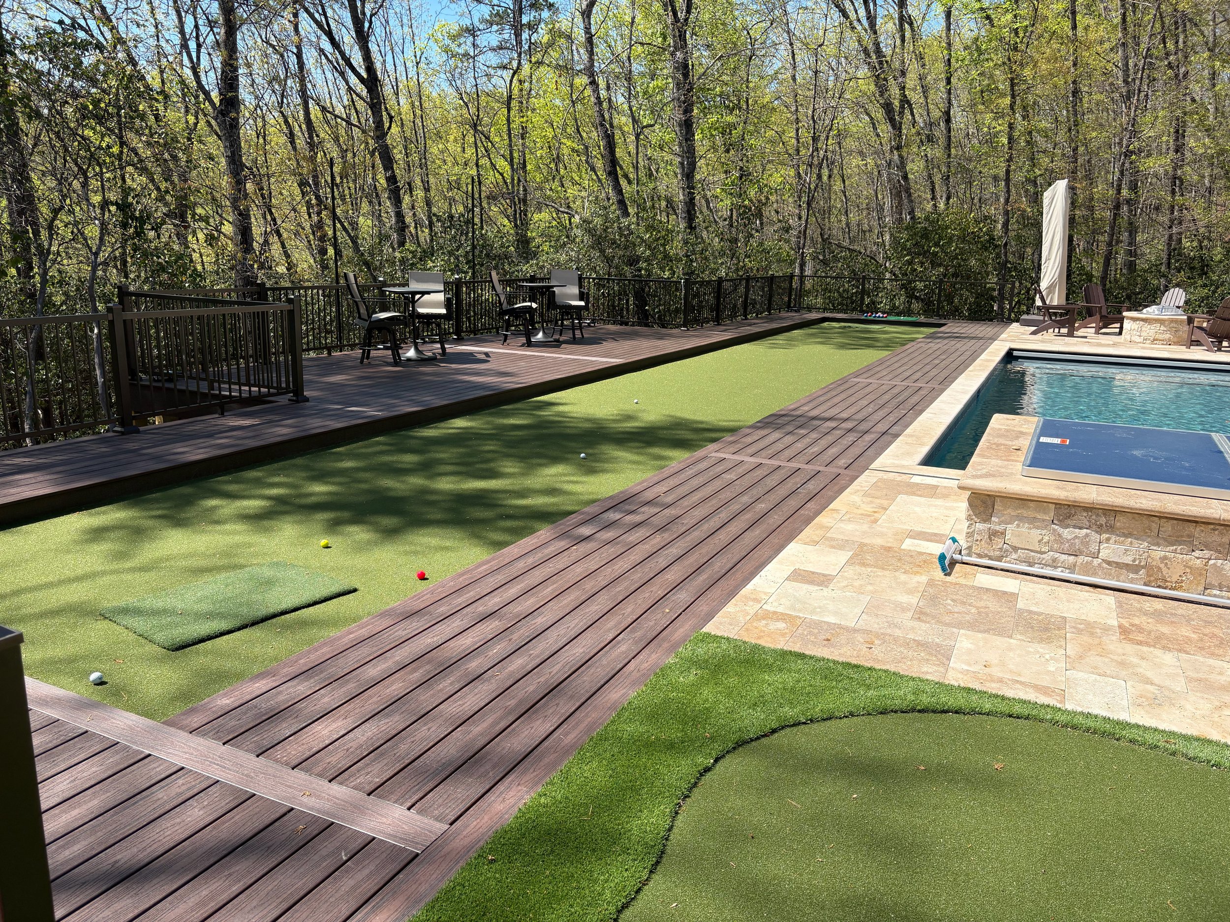 An outdoor patio area with a professional size bocce ball court, featuring green artificial turf and several scattered bocce balls, surrounded by a wooden deck and outdoor furniture, with a forested background.