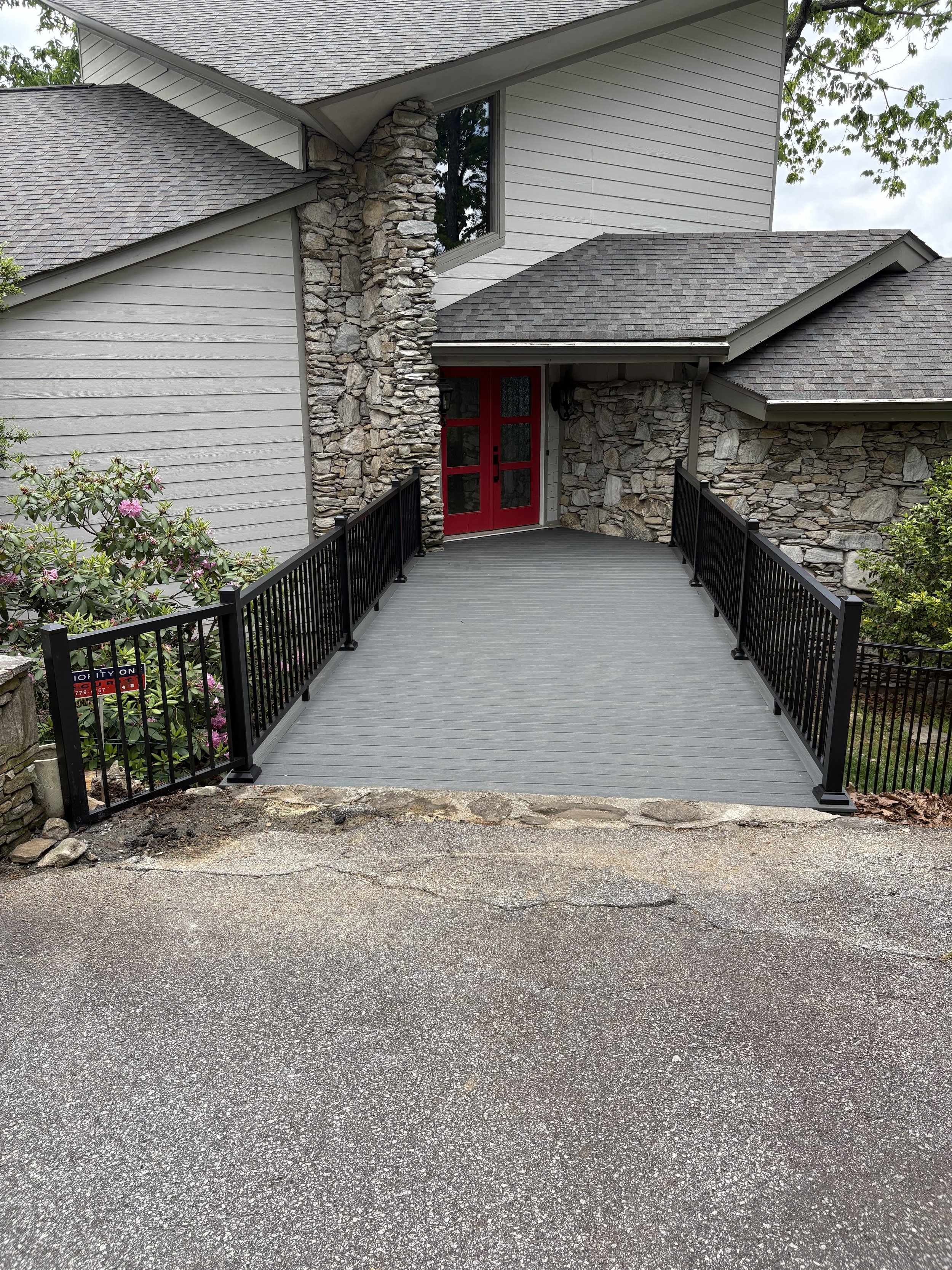 Entrance to a house with a red door, surrounded by stone and siding walls, with a wooden ramp leading up to it and a security system sign at the entrance.