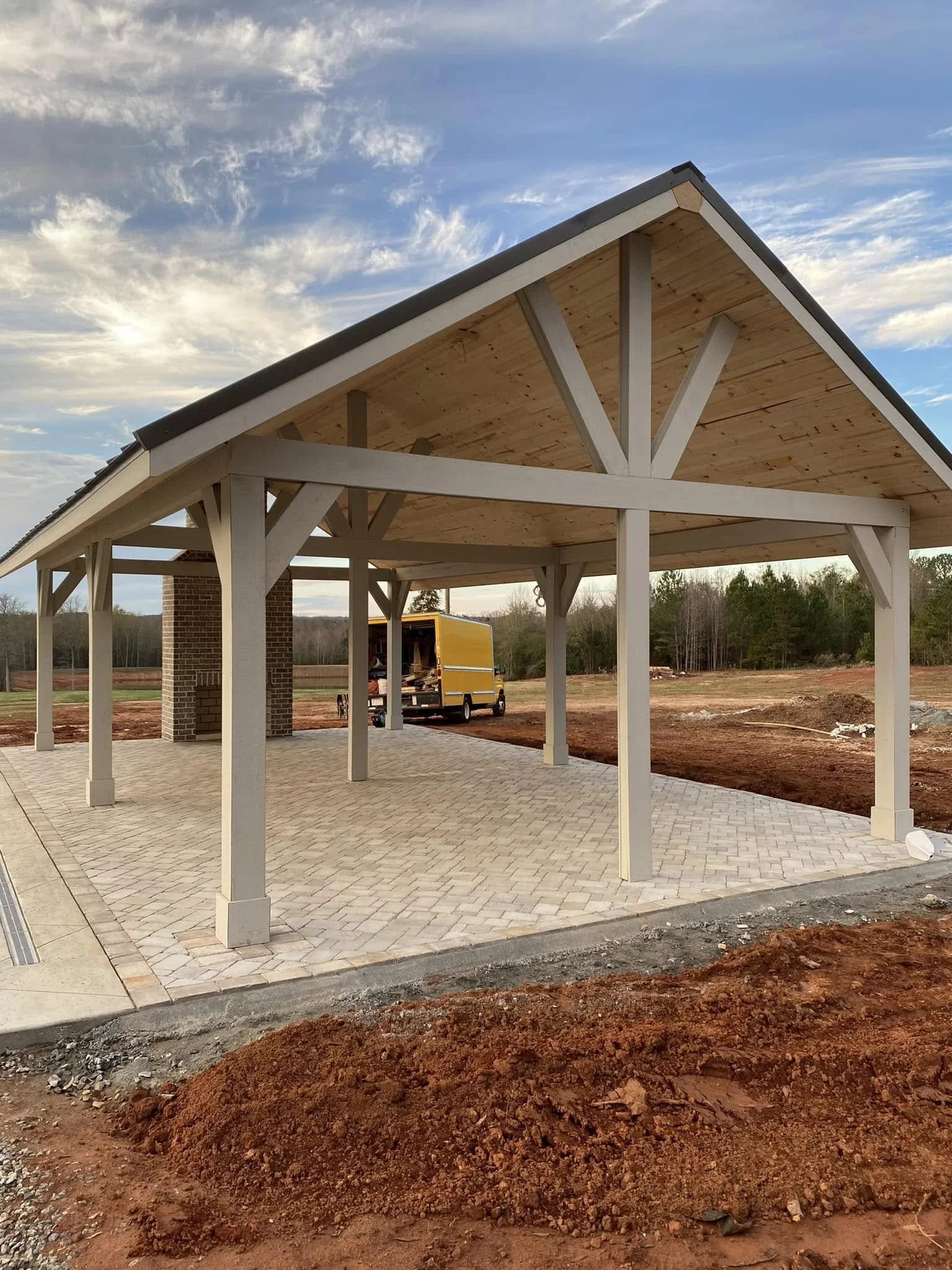 Under construction wooden pavilion with brick support columns on paved ground, background home and trees, cloudy sky.