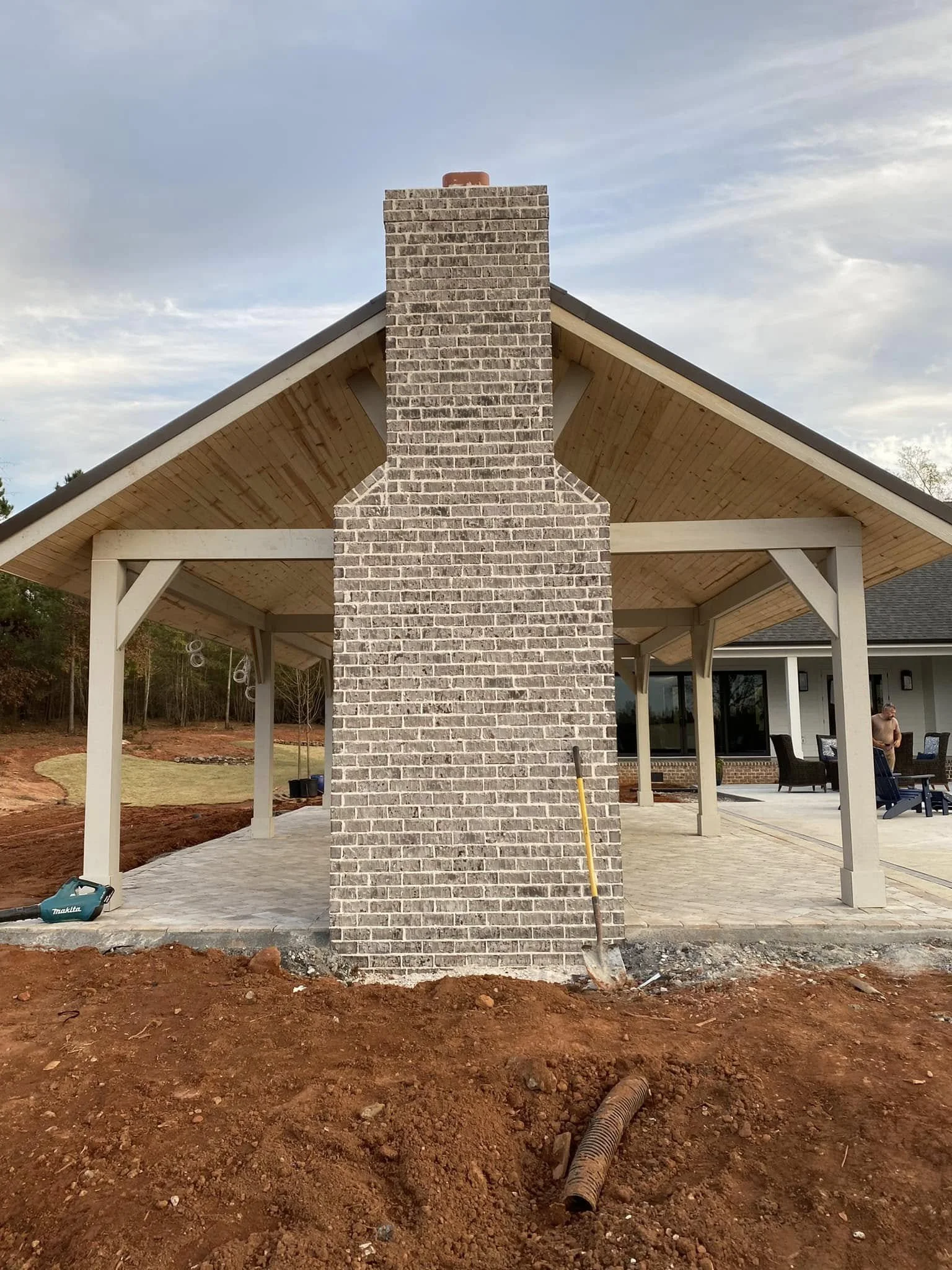 Backyard patio with brick chimney, covered seating area, and ongoing landscaping work with exposed soil and drainage pipe.