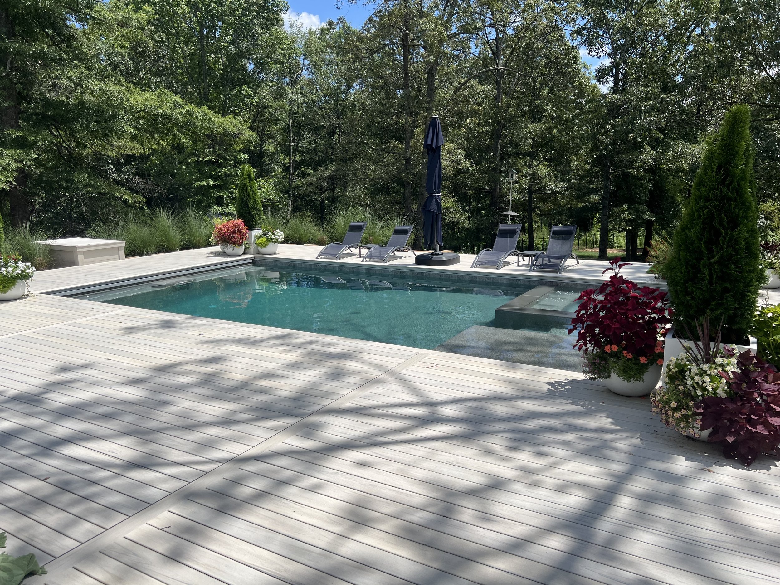 A backyard swimming pool surrounded by a white wooden deck with several lounge chairs, potted plants, and an outdoor umbrella, set against a backdrop of dense green trees.