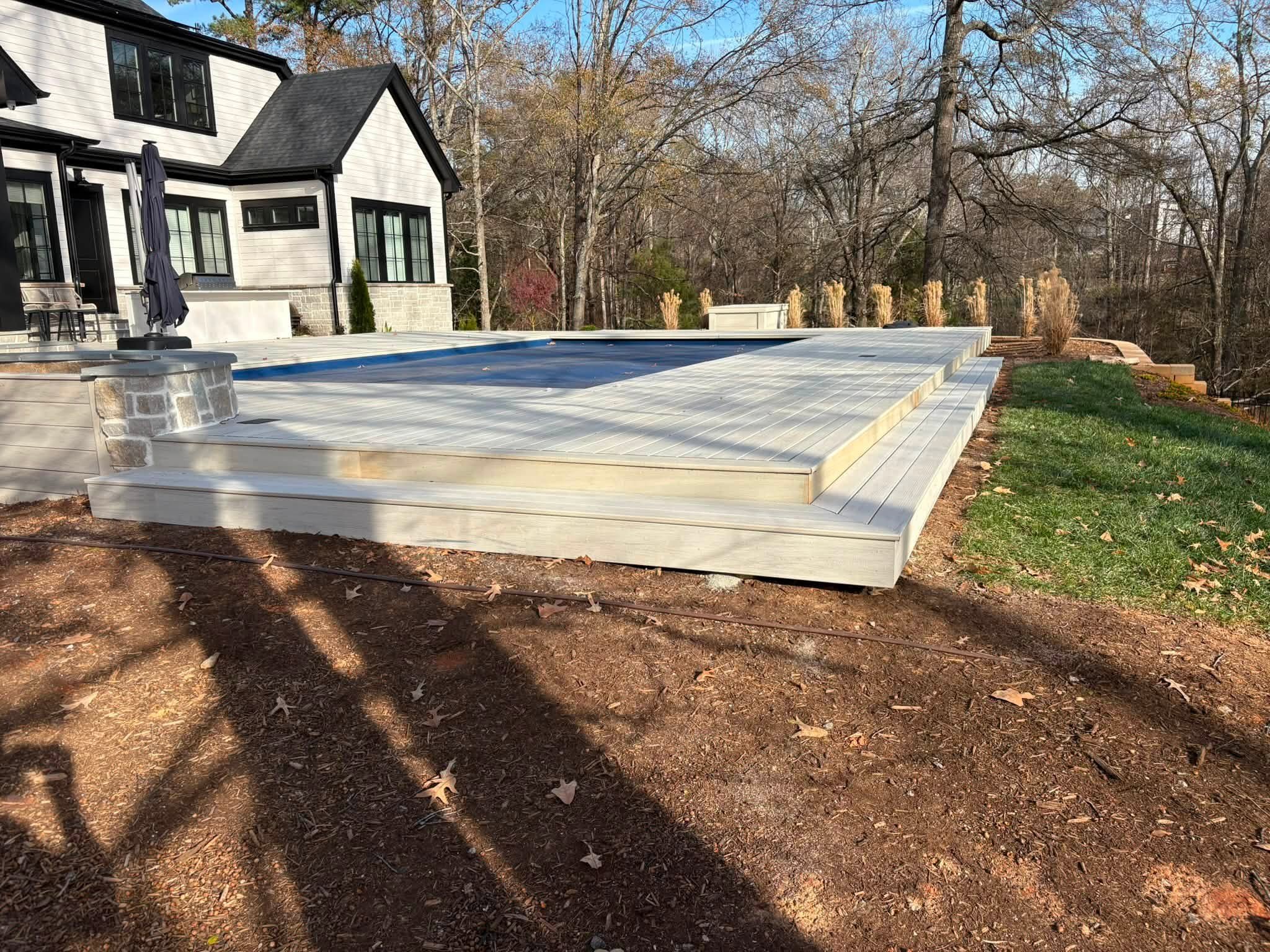 Backyard with a white house, a deck, and a swimming pool in a wooded area.