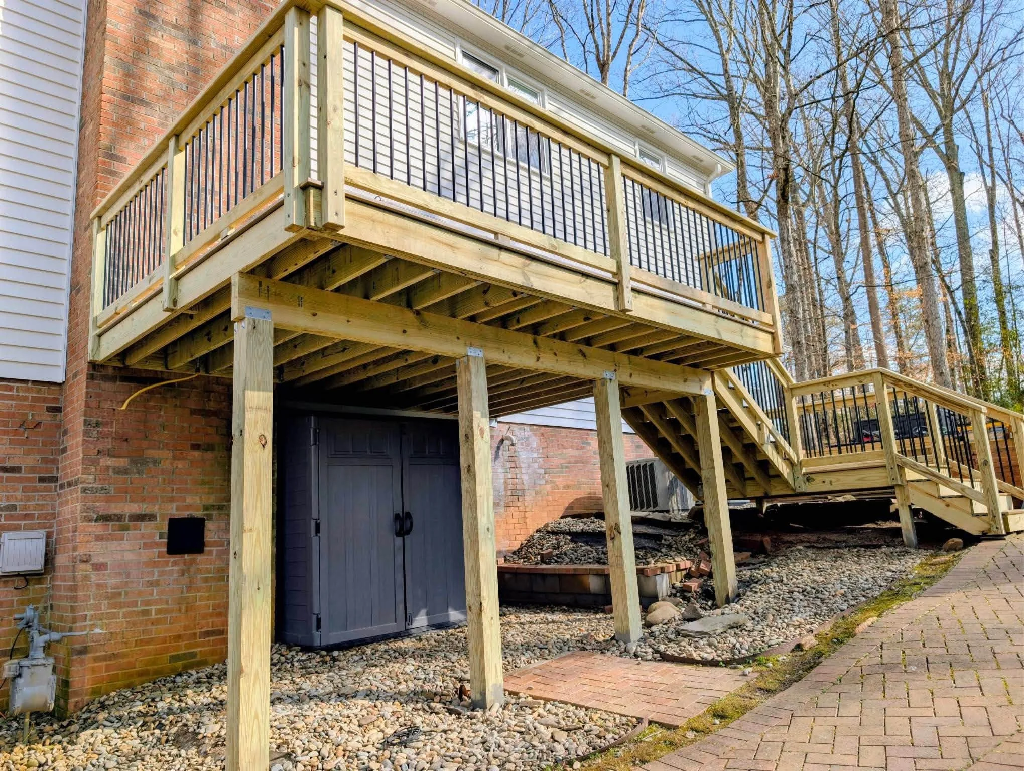 The image shows a new wooden deck being constructed outside a brick house. The deck has a railing with metal balusters and stairs leading down to the ground. The background features leafless trees and a clear blue sky.