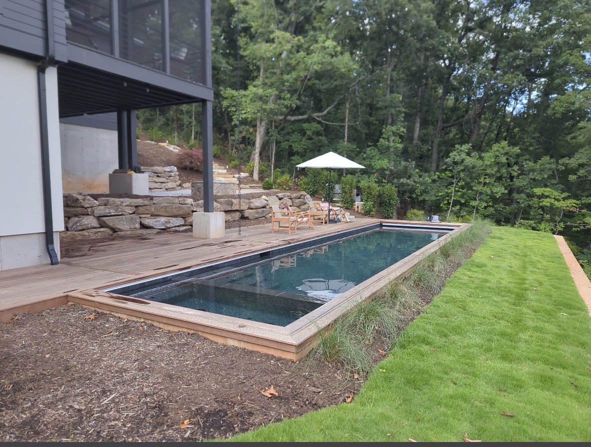 Backyard with a rectangular swimming pool, wooden deck, outdoor furniture, umbrella, and surrounded by trees and greenery.